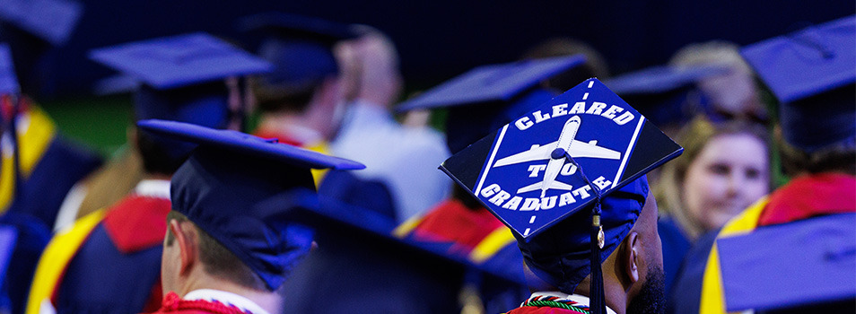 Graduates in blue caps and gowns wear mortarboards with a "Cleared to Graduate" airplane design, symbolizing their academic achievement.