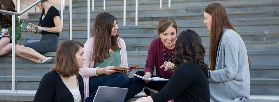 A group of young people sits on outdoor steps, engaged in conversation and study with laptops and notebooks.