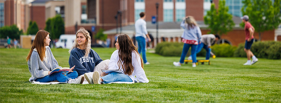 Three individuals sit on a blanket on a grassy field, engaged in conversation, with campus buildings and other people in the background.