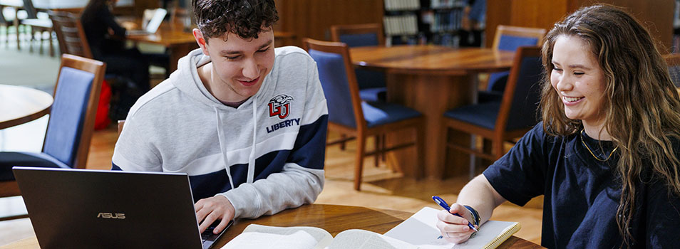Two students collaborate in a library, one using a laptop and the other taking notes in a notebook.