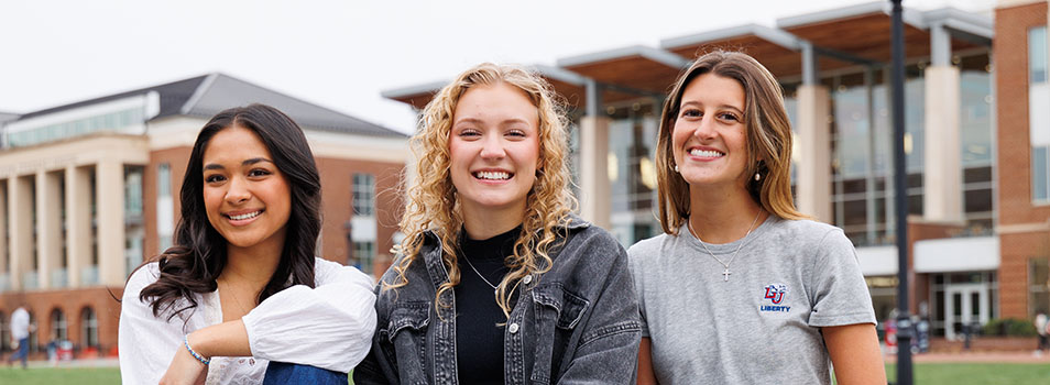 Three young individuals sit together on a bench outdoors in front of a university-style building, smiling for the camera.