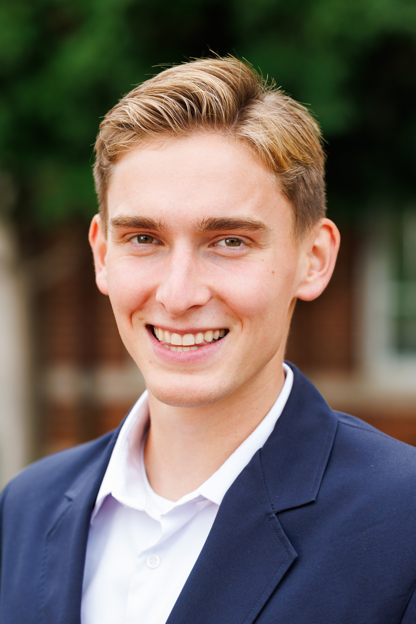 A young person with light brown hair and a smile wears a navy blazer over a white collared shirt, standing outdoors with greenery blurred in the background.