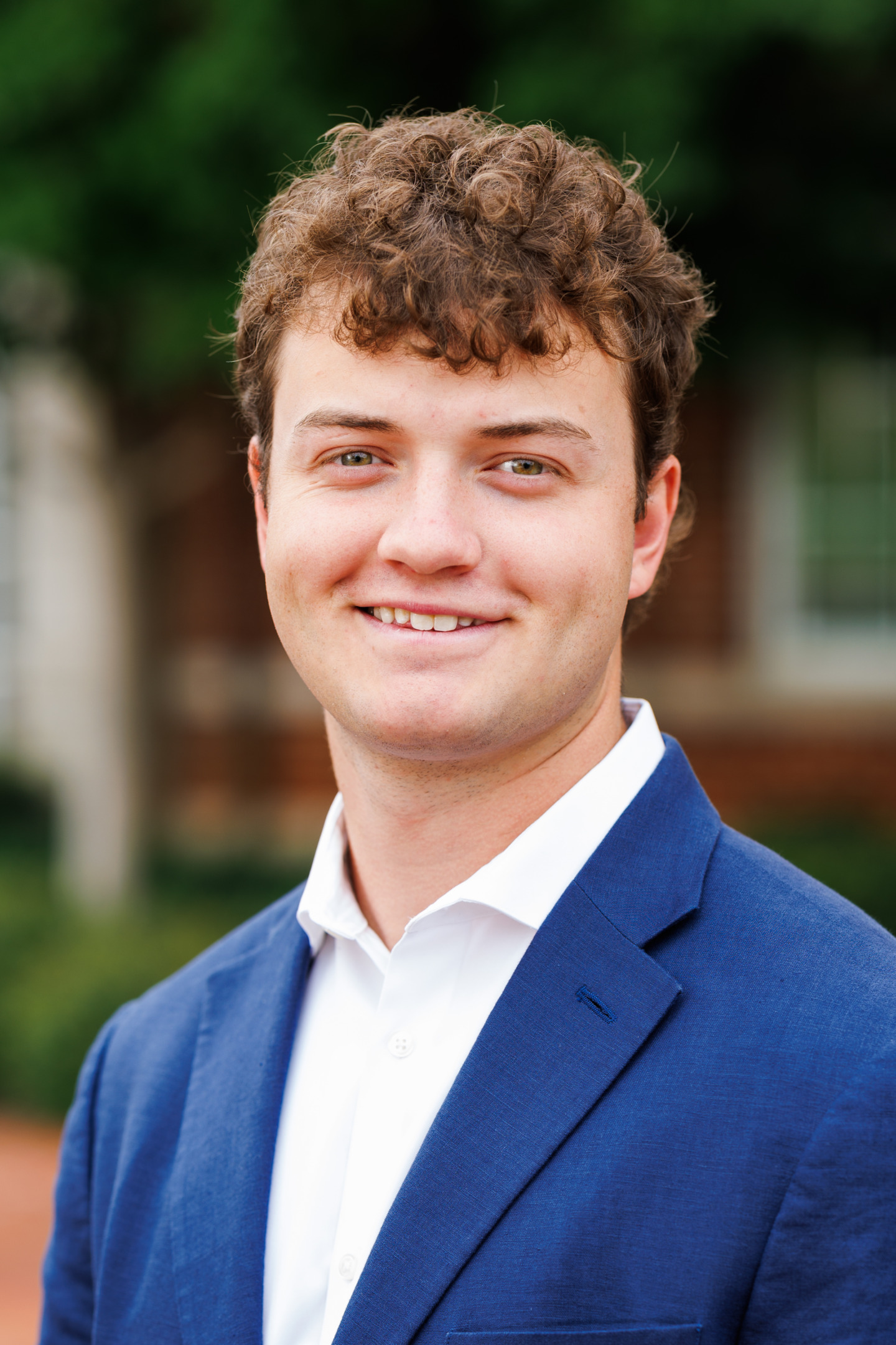 A young person with curly brown hair smiles warmly while wearing a blue blazer and white shirt outdoors.