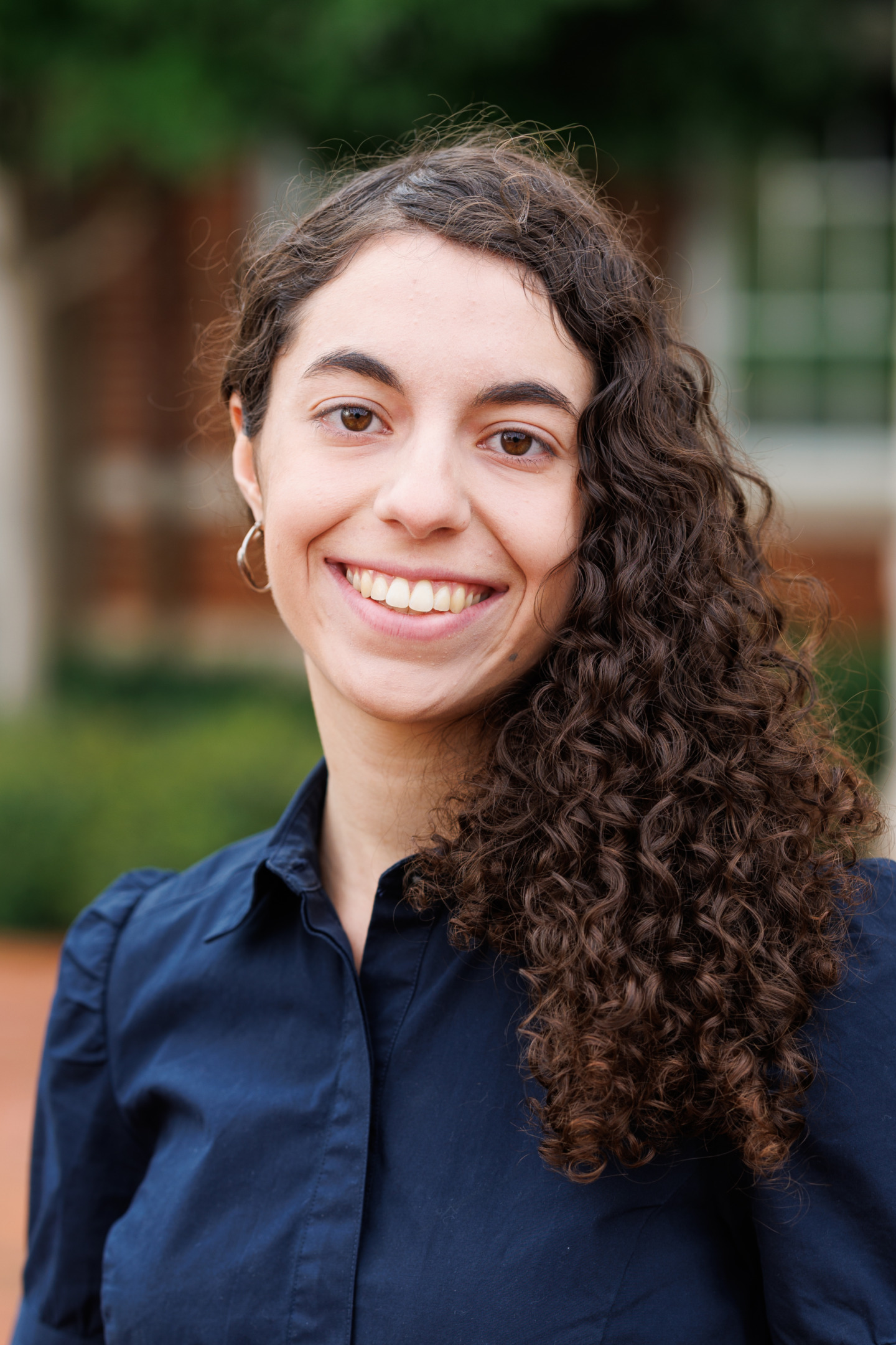 A person with long curly brown hair and a bright smile wears a dark blue shirt outdoors.
