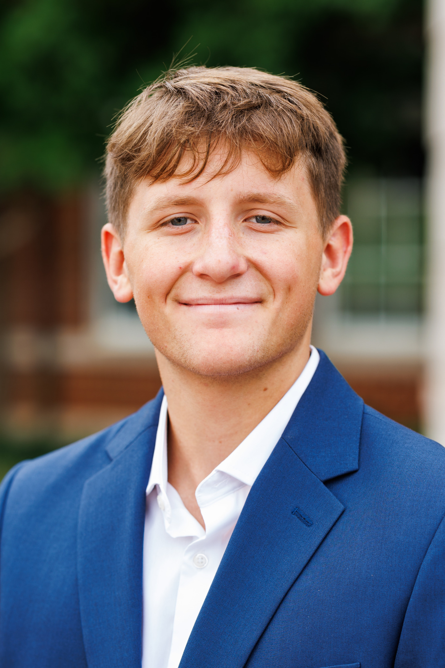 A young person with light brown hair smiles warmly while wearing a blue blazer over a white collared shirt, set against a softly blurred outdoor background.