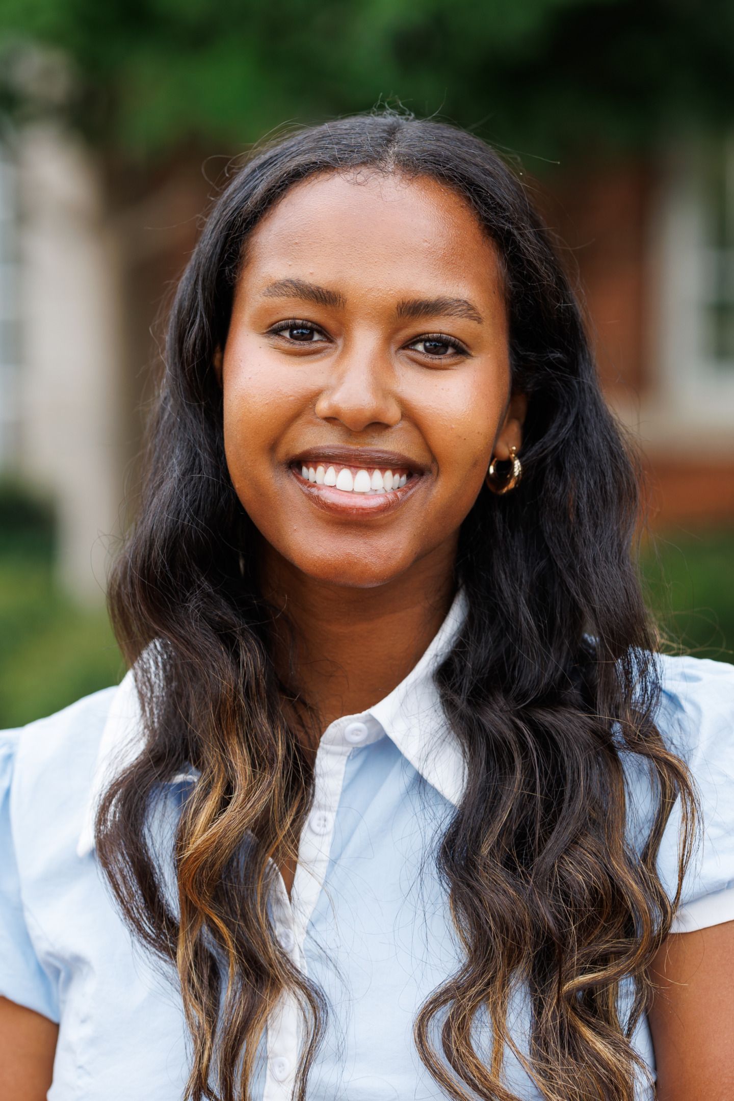 A smiling person with long, wavy dark hair and a light blue collared shirt stands outdoors against a blurred background of greenery and buildings.