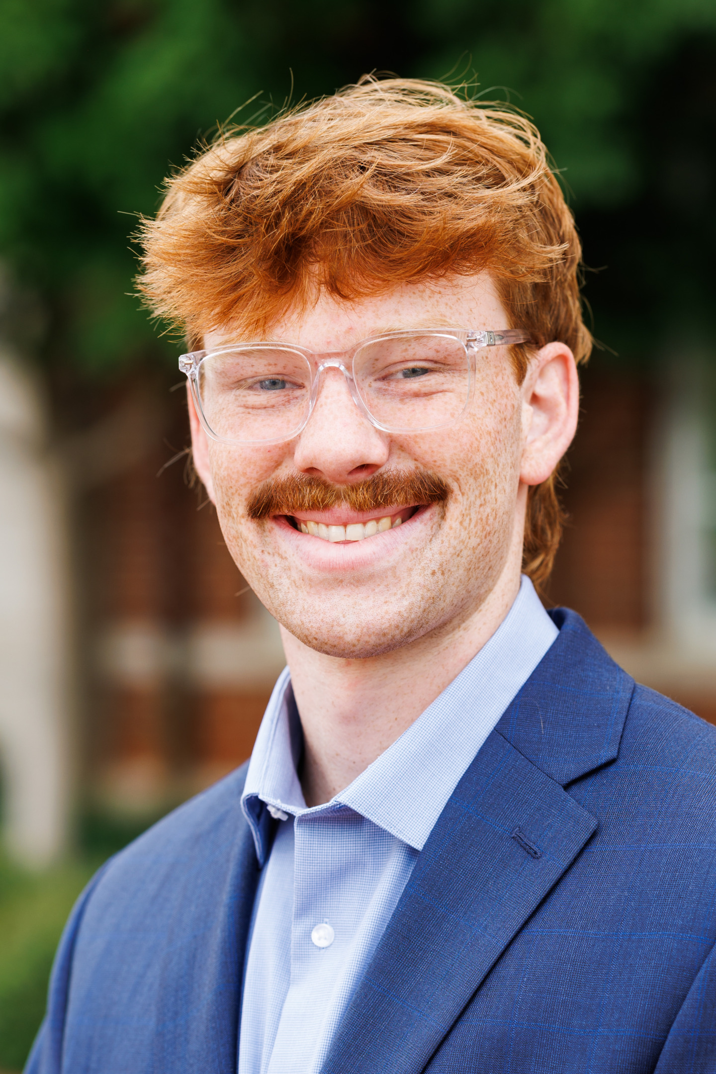 A person with red hair and a mustache, wearing clear glasses and a blue suit, smiles warmly at the camera.