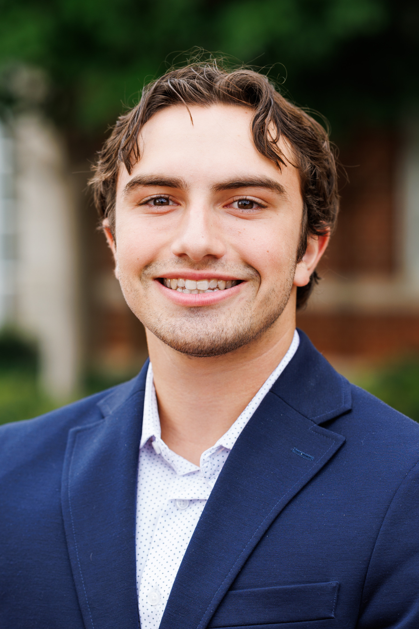 A young person with dark, wavy hair smiles warmly while wearing a navy blazer over a light-colored patterned shirt, set against a softly blurred outdoor background.
