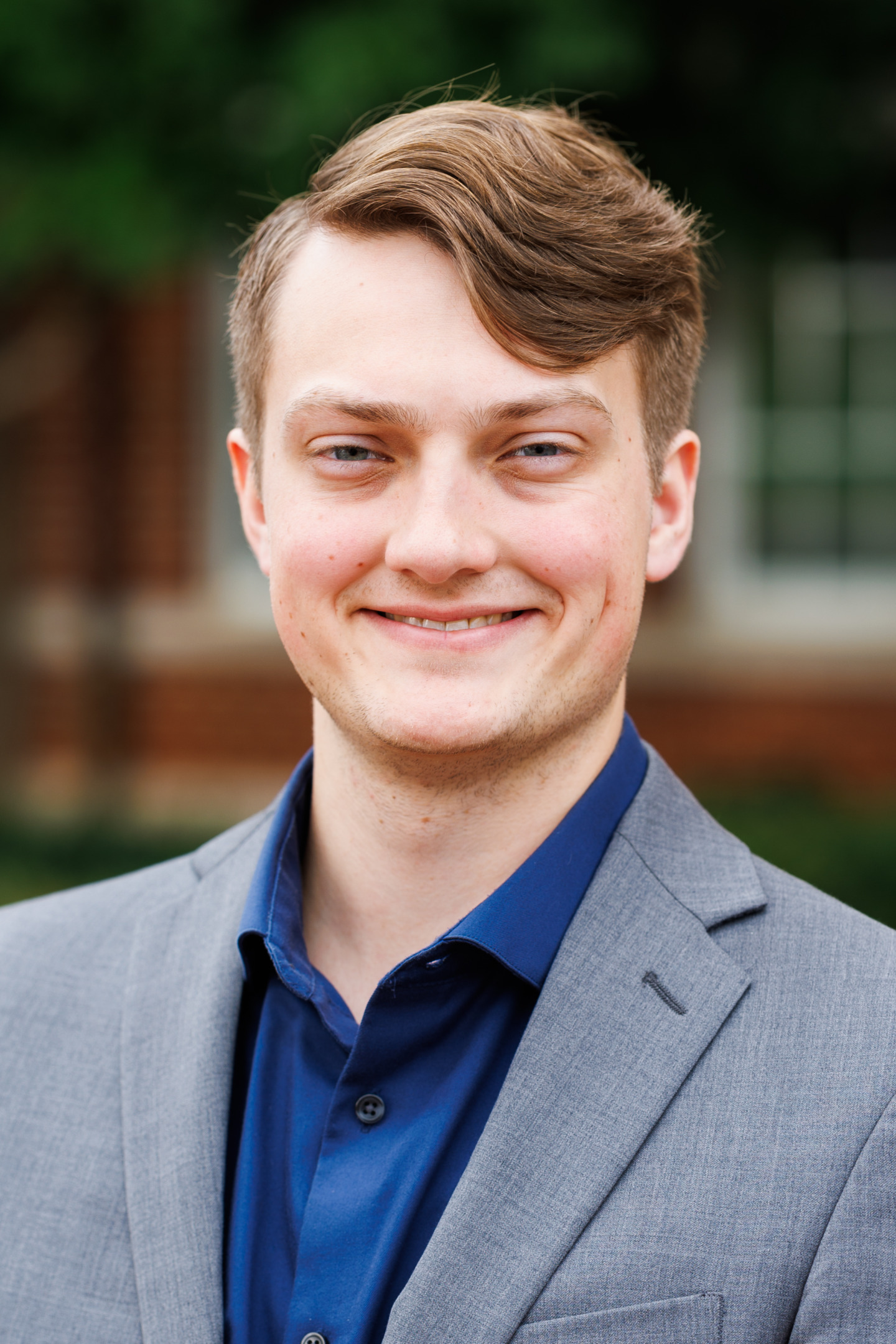 A young person with light brown hair and blue eyes smiles warmly while wearing a gray blazer over a blue collared shirt, standing outdoors against a blurred background of greenery and brick.