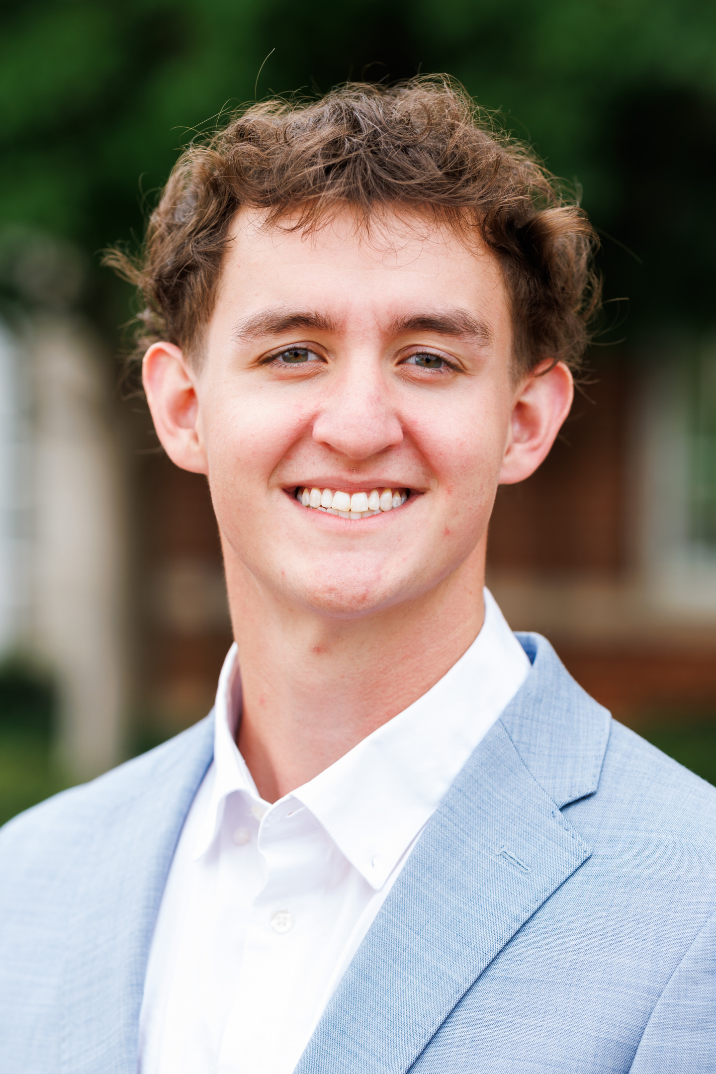 A young person with curly brown hair smiles warmly while wearing a light blue blazer over a white collared shirt, set against a softly blurred outdoor background.