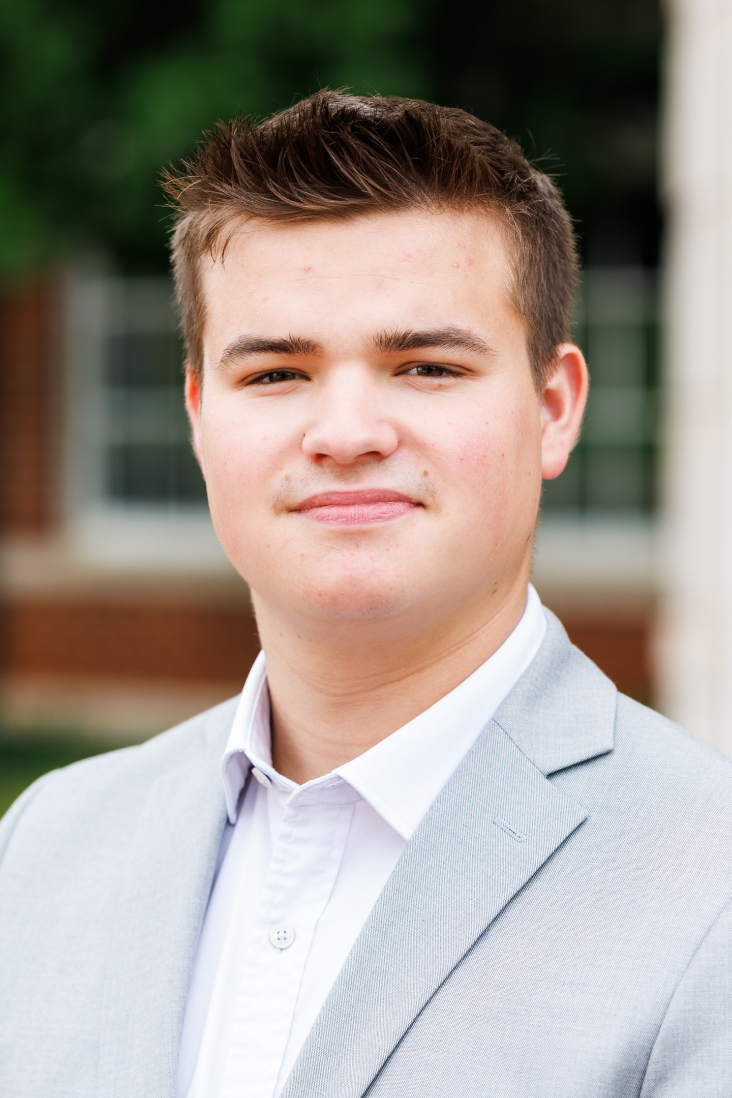A young person with short brown hair wears a light gray suit jacket over a white collared shirt and smiles slightly while looking at the camera.