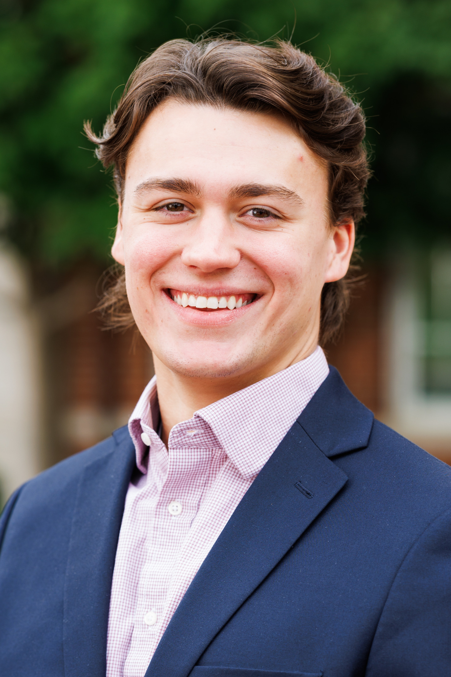 A smiling young person with wavy brown hair wears a navy blazer over a pink and white checkered shirt, standing outdoors with blurred greenery in the background.