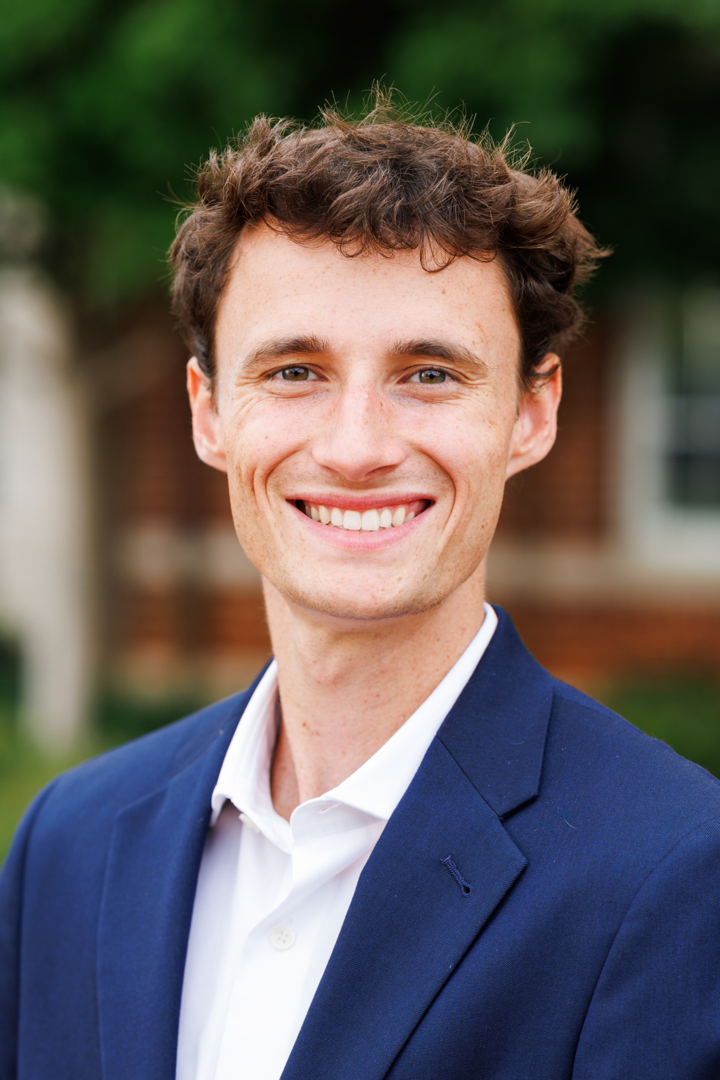 A smiling young person with curly brown hair wears a navy blazer over a white shirt, standing outdoors with blurred greenery and brick buildings in the background.