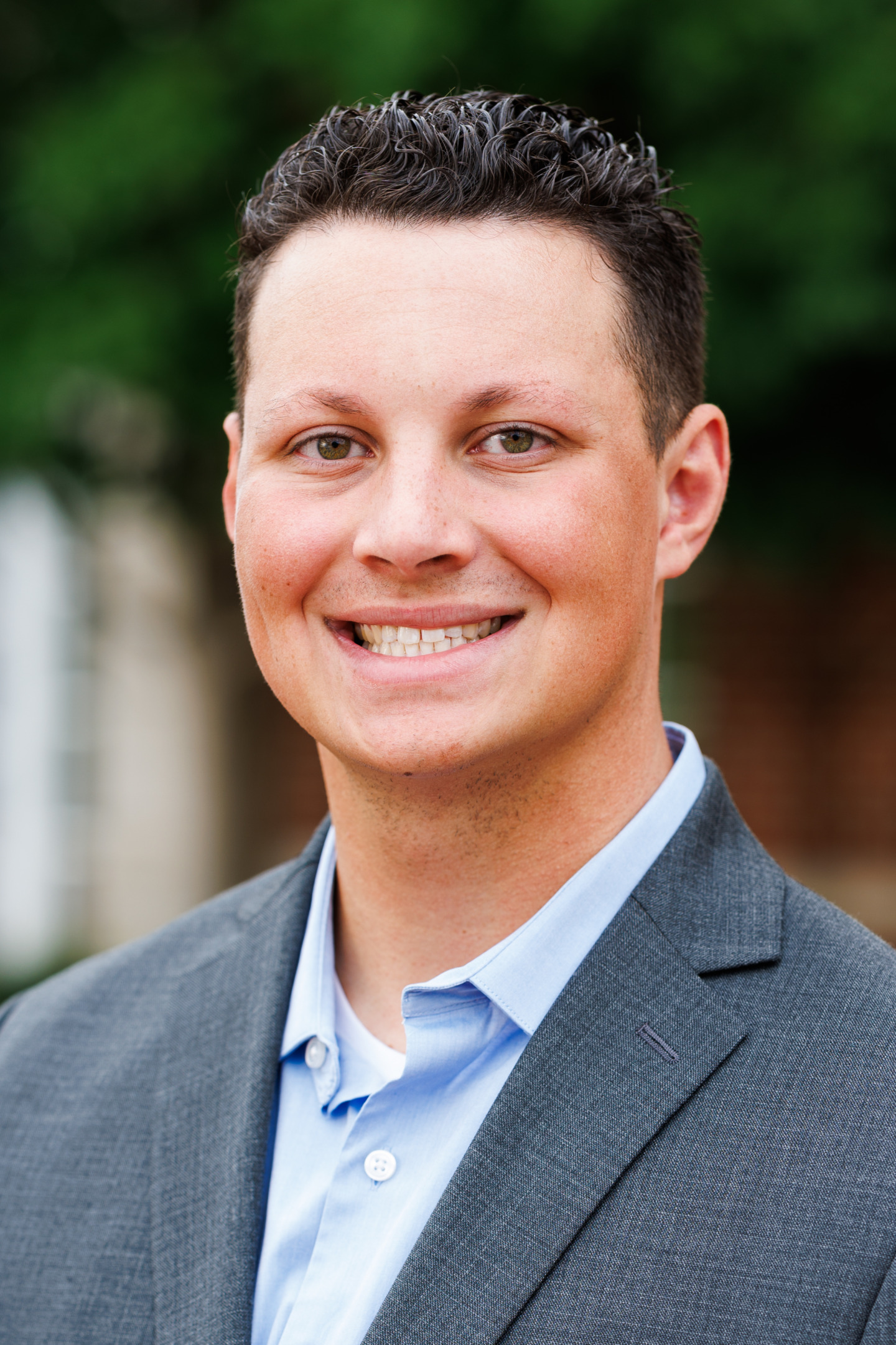 A smiling person with short dark hair wears a gray blazer over a light blue collared shirt, standing outdoors with greenery in the background.