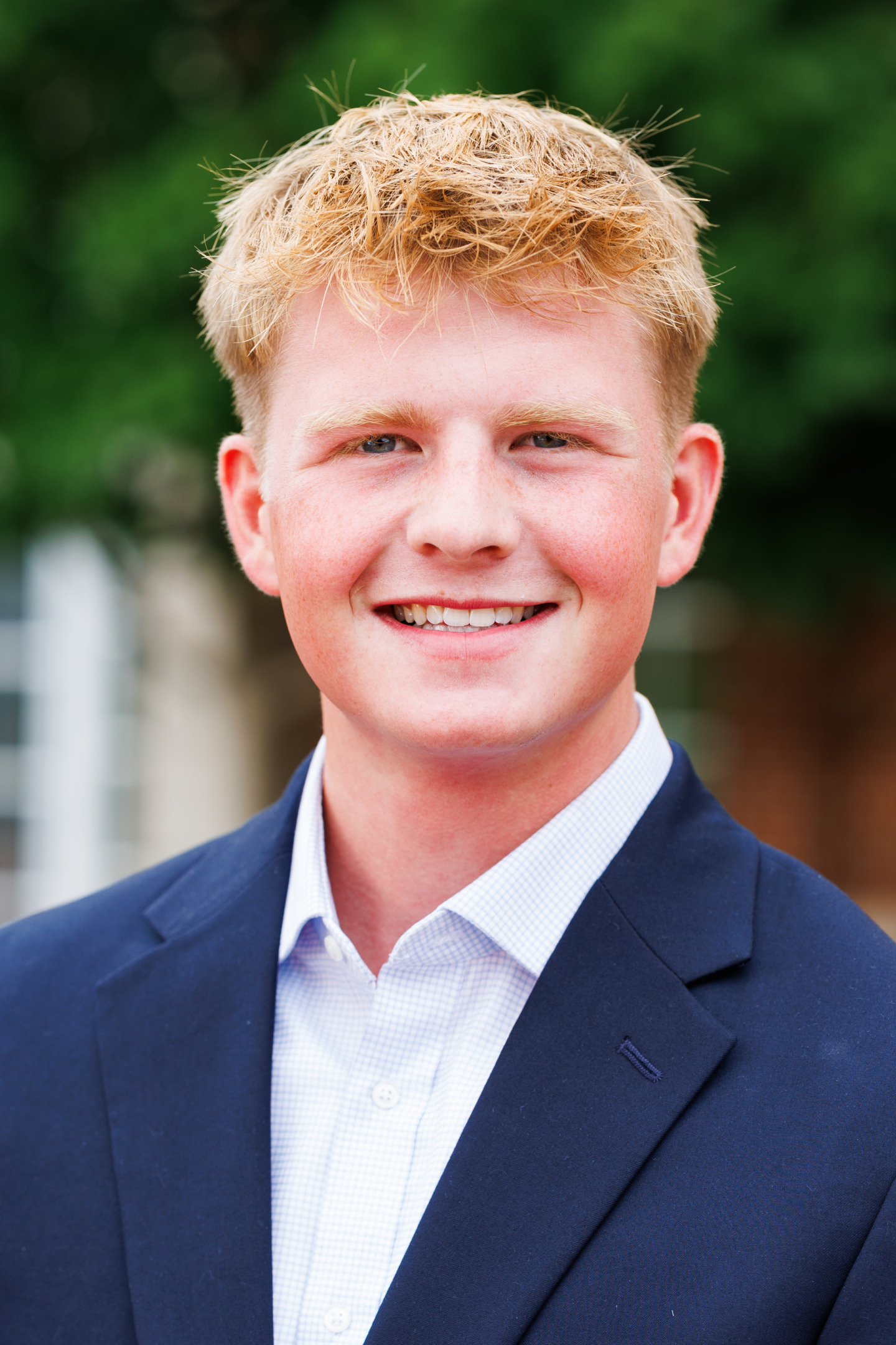A young person with blond hair smiles warmly while wearing a navy blazer over a light-colored collared shirt, set against a blurred green outdoor background.