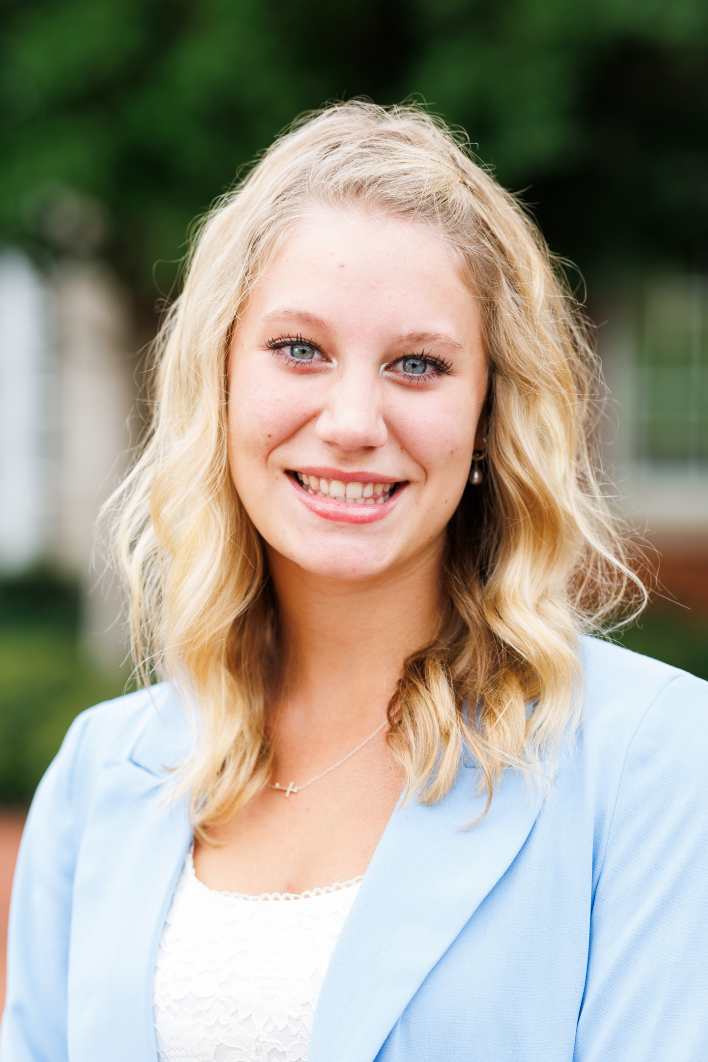 A smiling person with blonde wavy hair wears a light blue blazer over a white top, standing outdoors with a blurred green background.