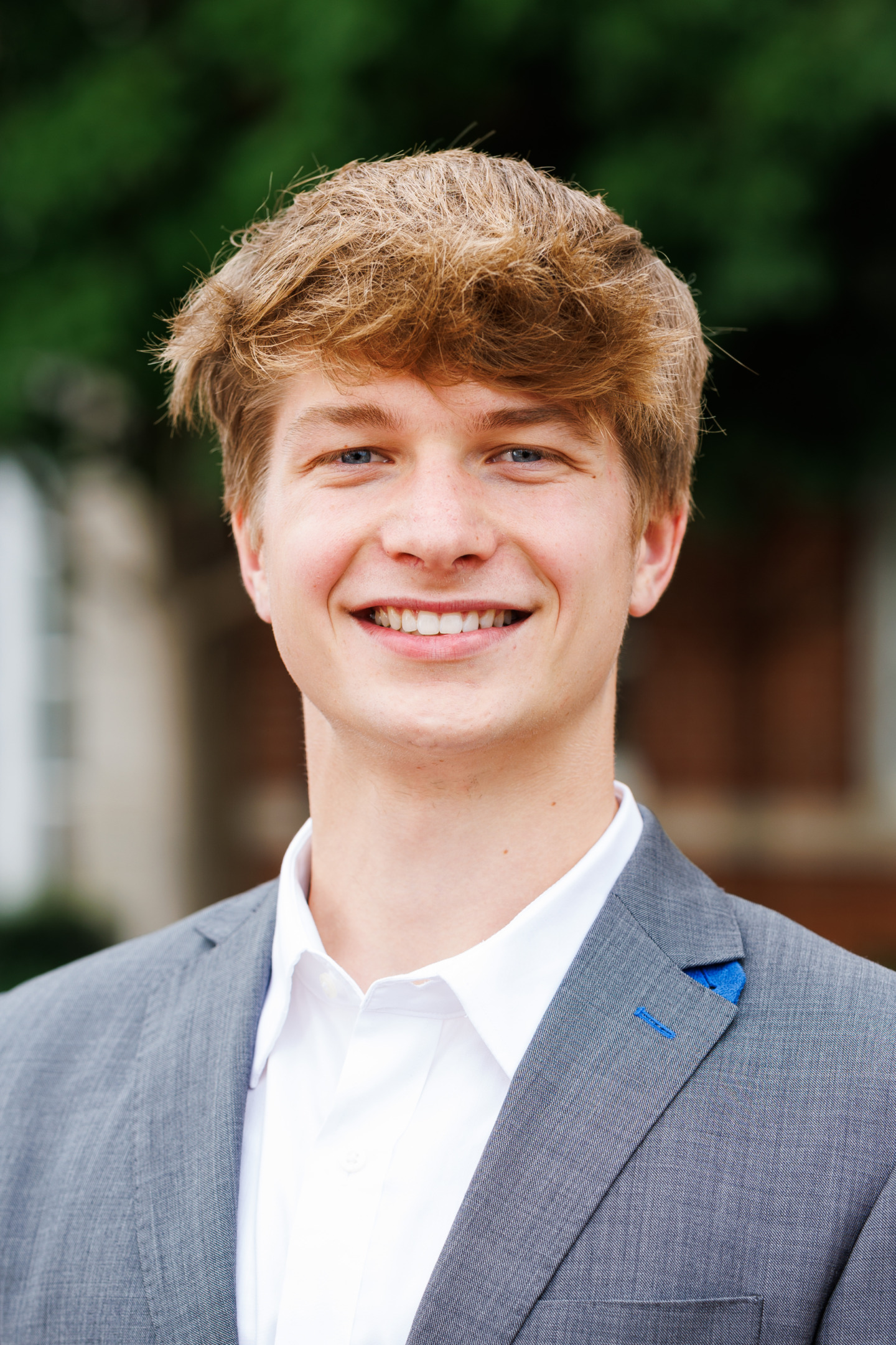 A young person with light brown hair and blue eyes smiles warmly while wearing a gray suit jacket over a white shirt, standing outdoors with green foliage in the background.
