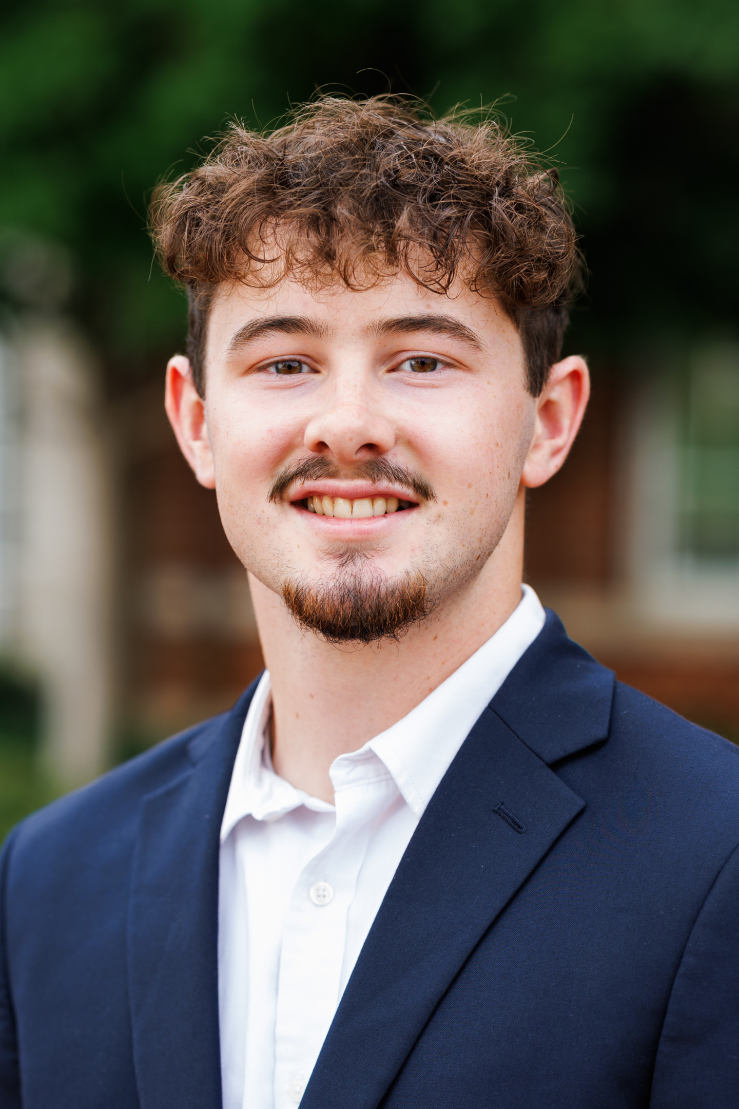 A young person with curly brown hair and a goatee smiles warmly while wearing a dark suit jacket and white shirt, set against a softly blurred outdoor background.