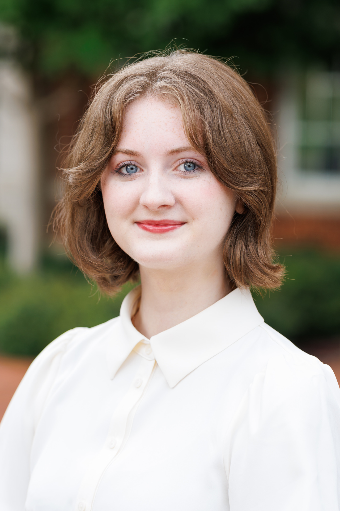 A young person with short brown hair and blue eyes smiles gently while wearing a white collared shirt outdoors.
