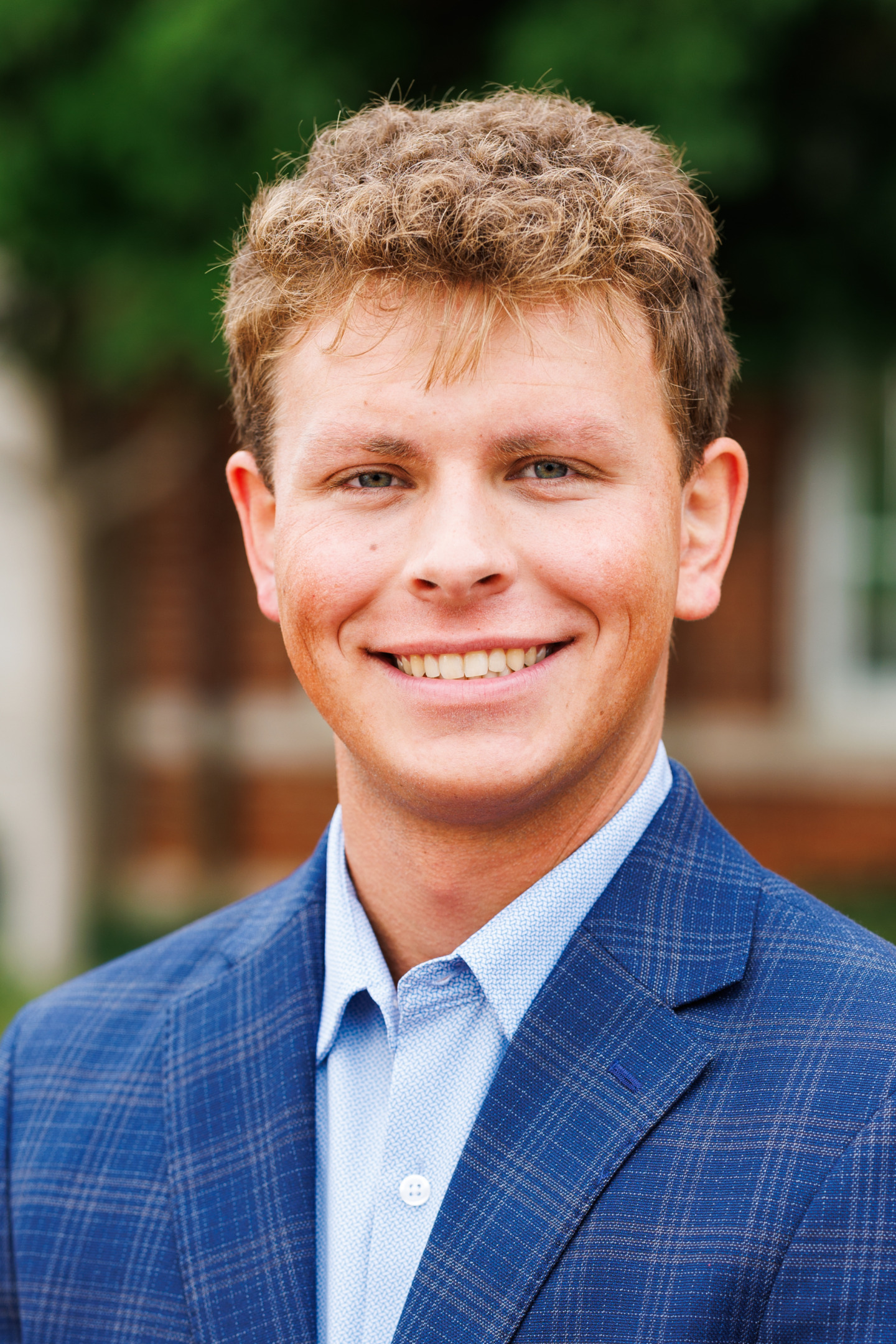 A young person with curly light brown hair smiles warmly while wearing a blue plaid blazer over a light blue collared shirt, set against a softly blurred outdoor background.
