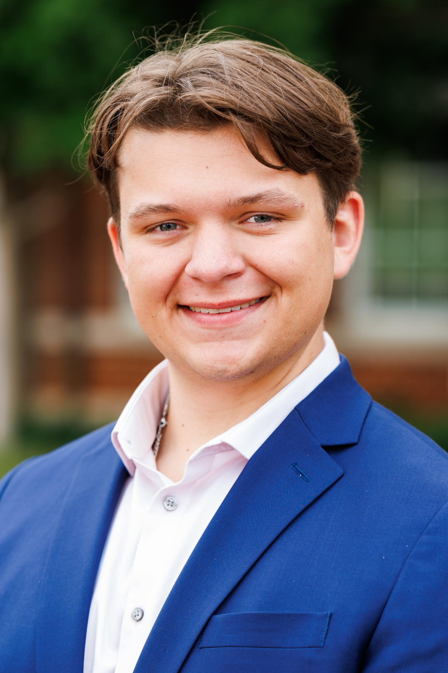 A young person with light brown hair smiles warmly while wearing a blue blazer and a light pink collared shirt.