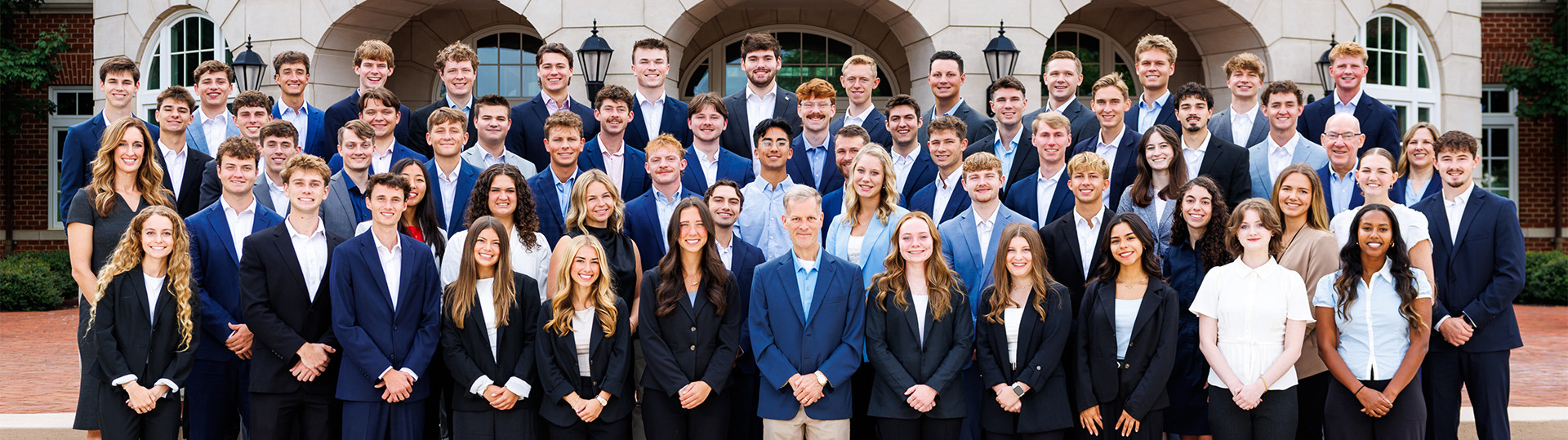 A group of young people in formal attire pose together on steps outside a stone building with arched windows.