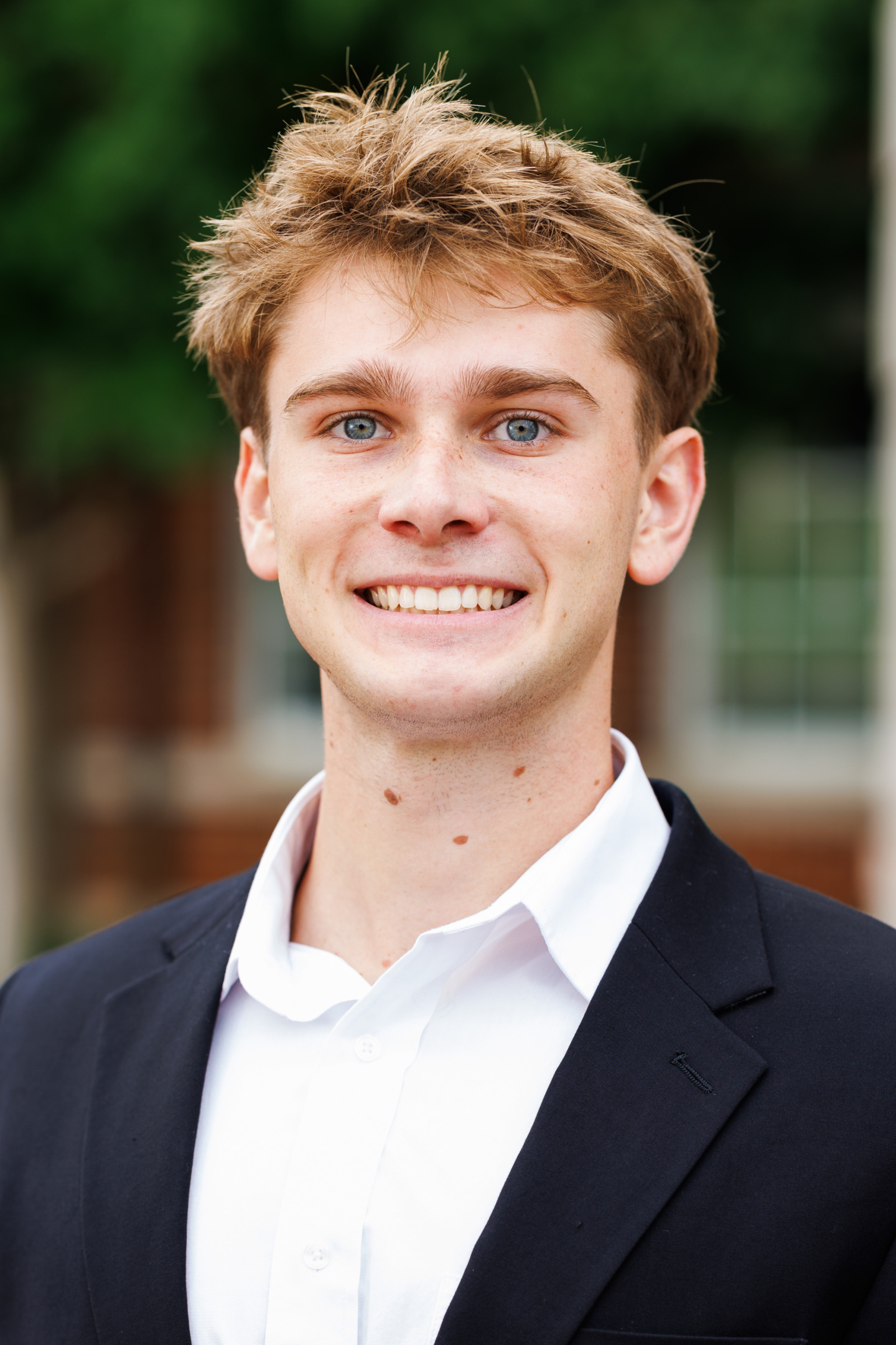 A young person with light brown hair and blue eyes smiles warmly while wearing a dark suit jacket over a white collared shirt, set against a softly blurred outdoor background.