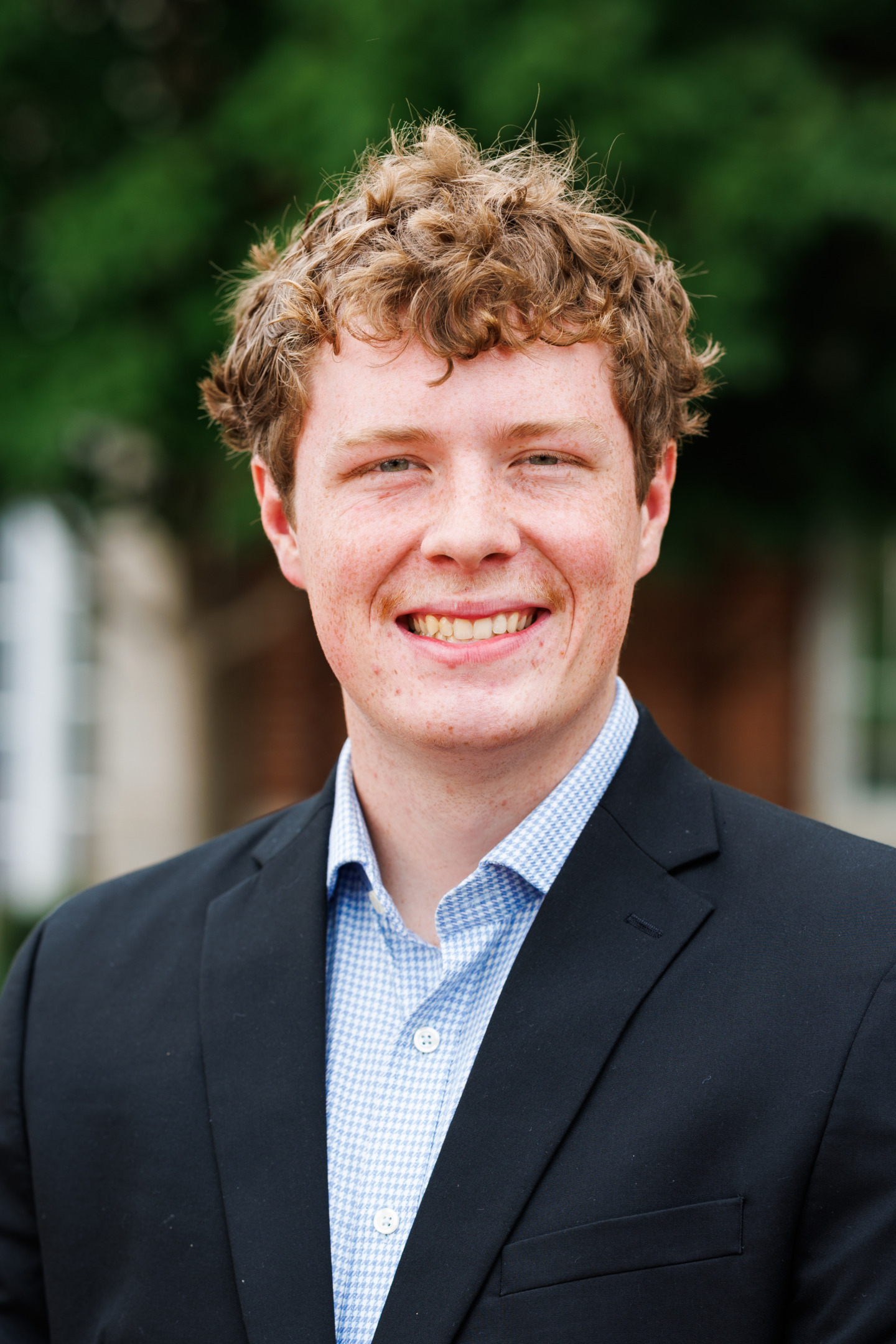A young person with curly hair smiles warmly while wearing a dark suit jacket over a light blue patterned shirt, standing outdoors with greenery in the background.