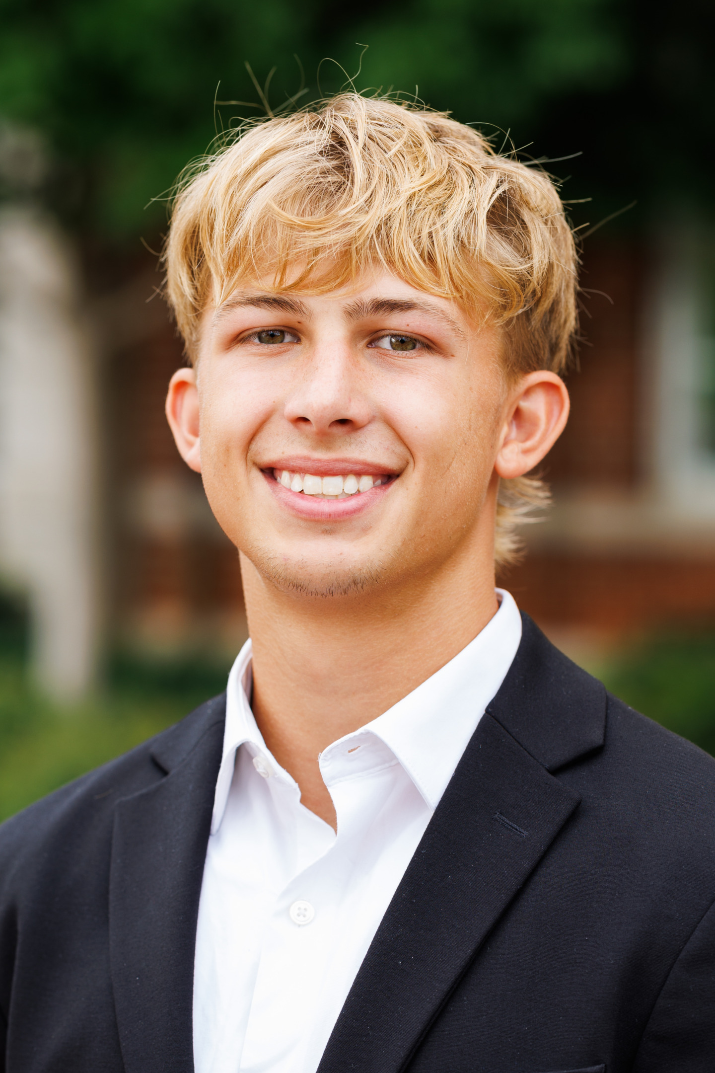 A young person with blonde hair smiles warmly while wearing a dark suit jacket and a white collared shirt, set against a softly blurred outdoor background.