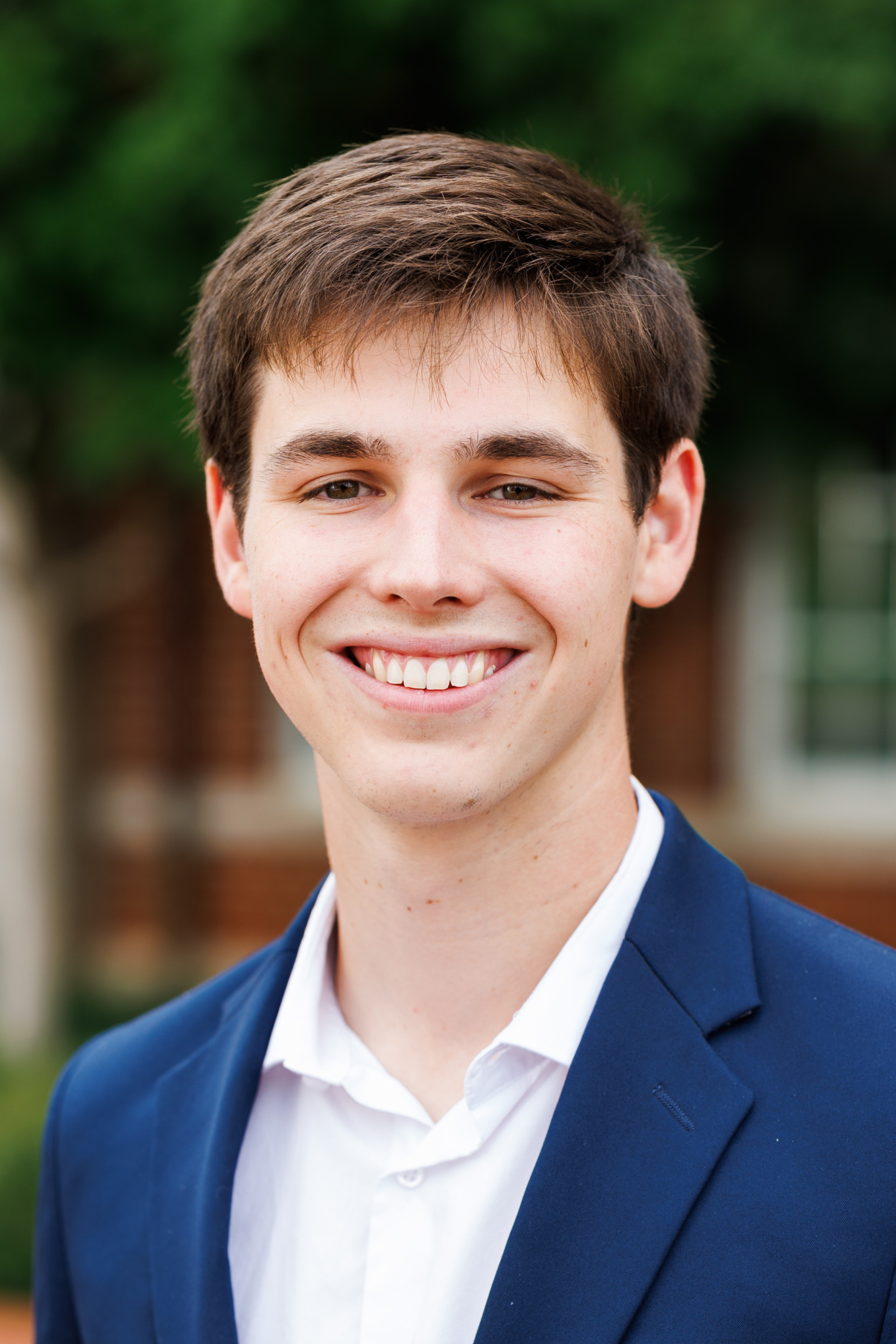 A young person with short brown hair smiles warmly while wearing a navy blue blazer over a white collared shirt, set against a blurred outdoor background.