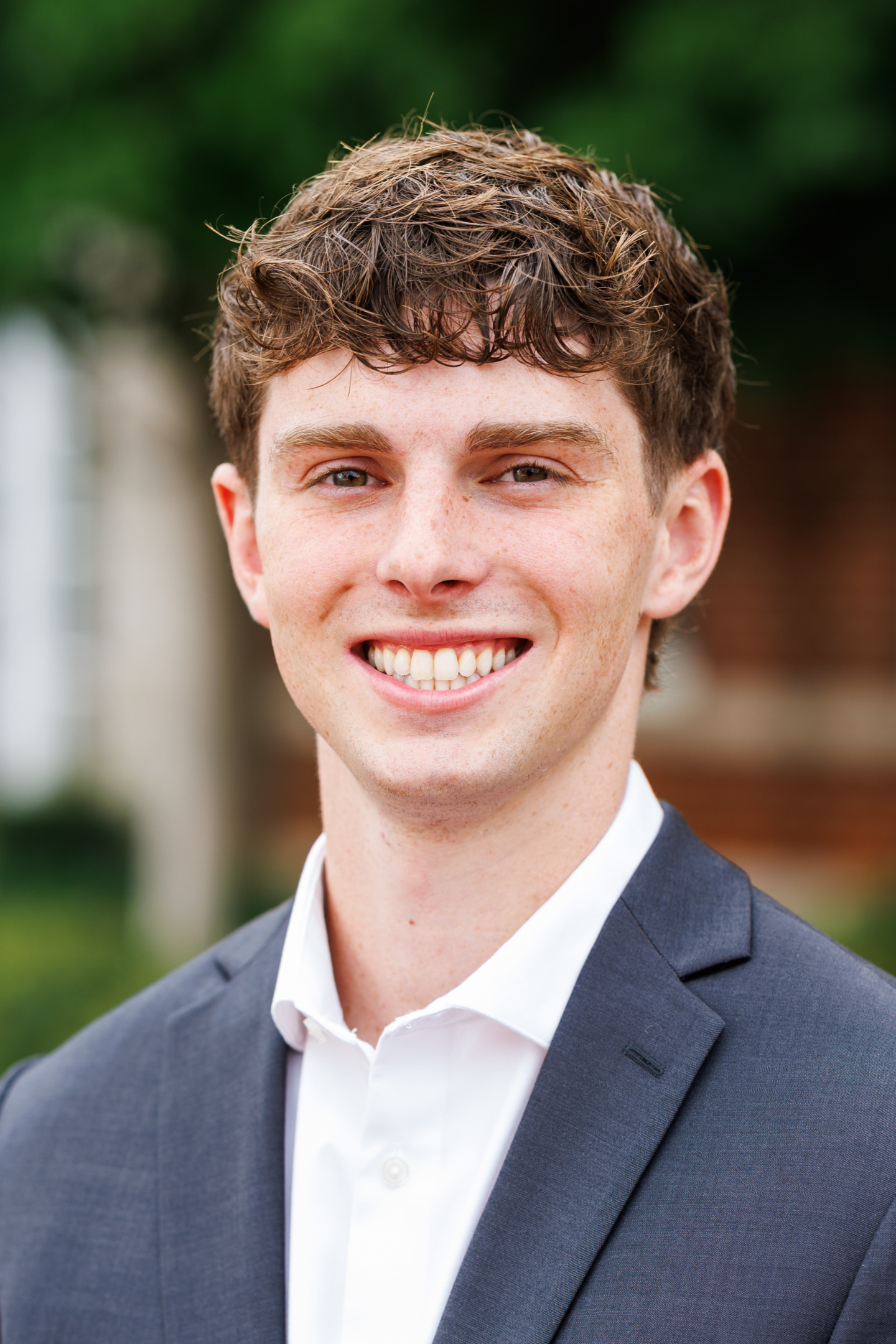 A young person with curly brown hair smiles warmly while wearing a dark suit jacket and a white collared shirt, set against a blurred outdoor background.
