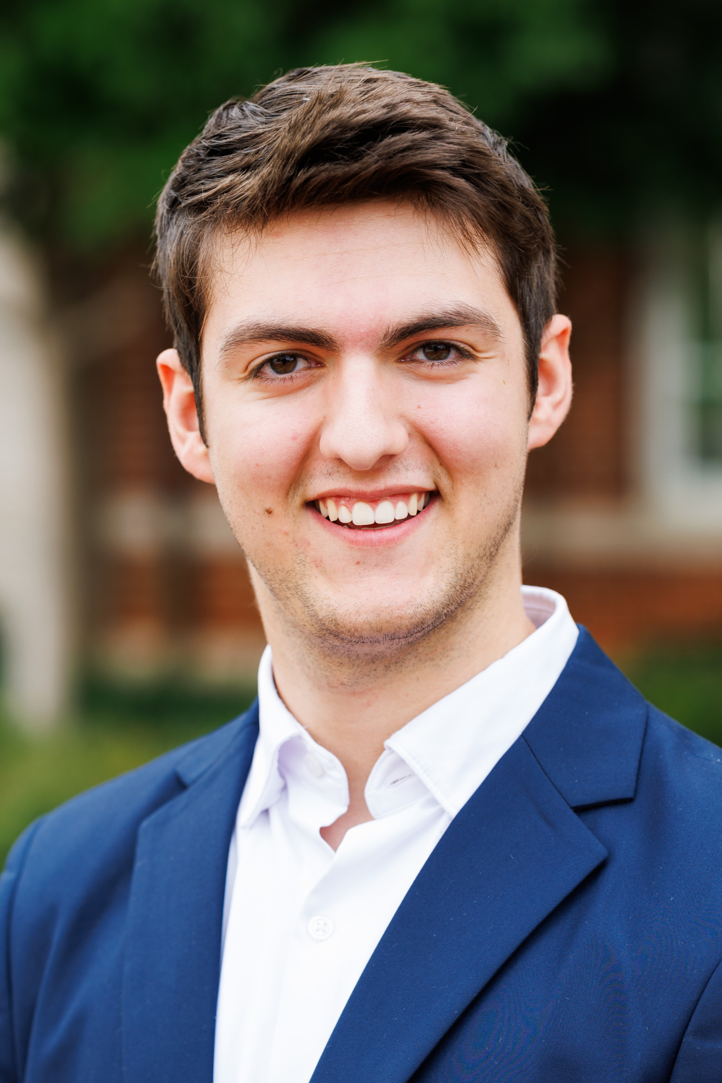 A young person with short brown hair smiles warmly while wearing a navy blazer over a white collared shirt, set against a blurred outdoor background.