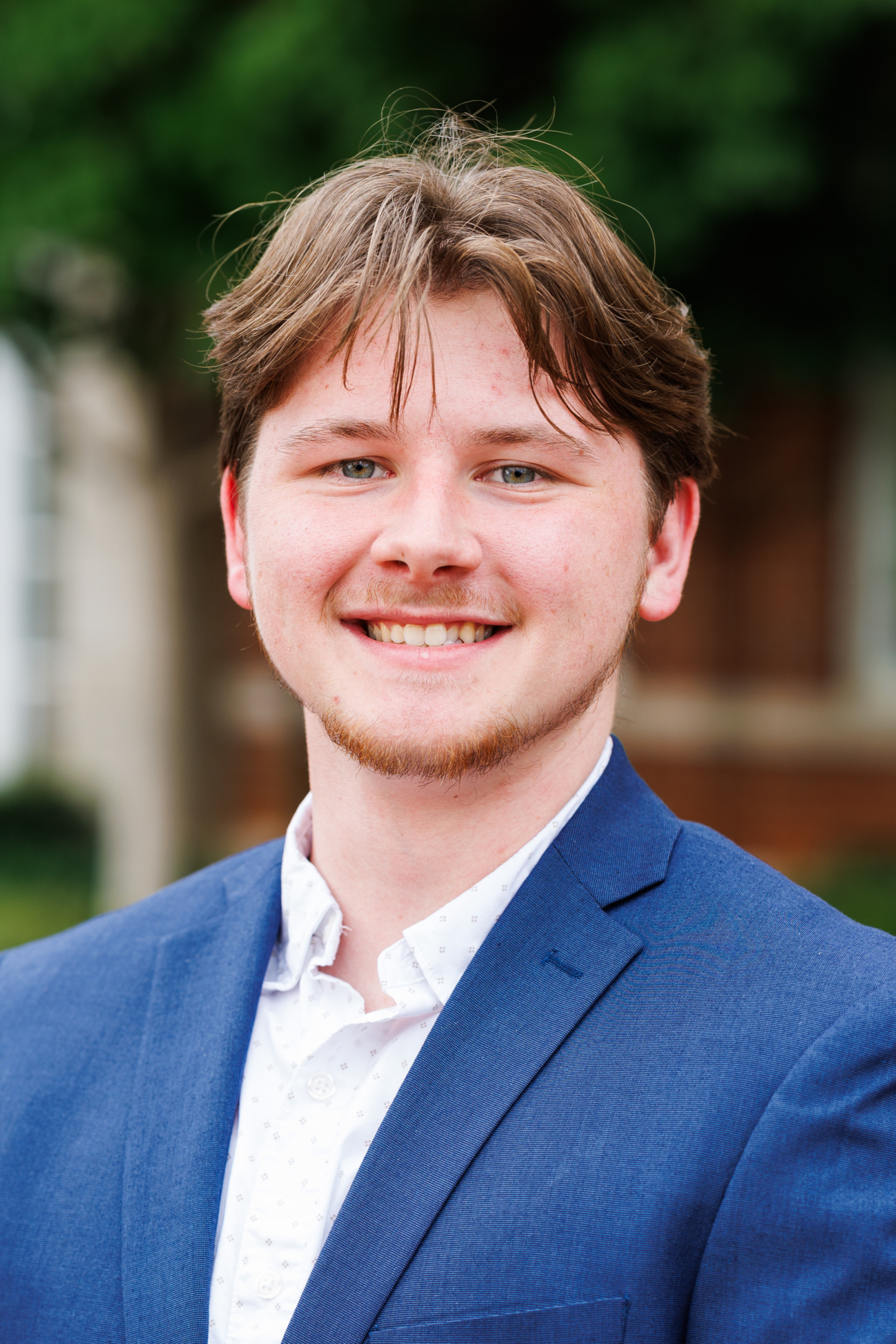 A young person with light brown hair and a beard smiles warmly while wearing a blue blazer over a white patterned shirt, set against a blurred outdoor background.