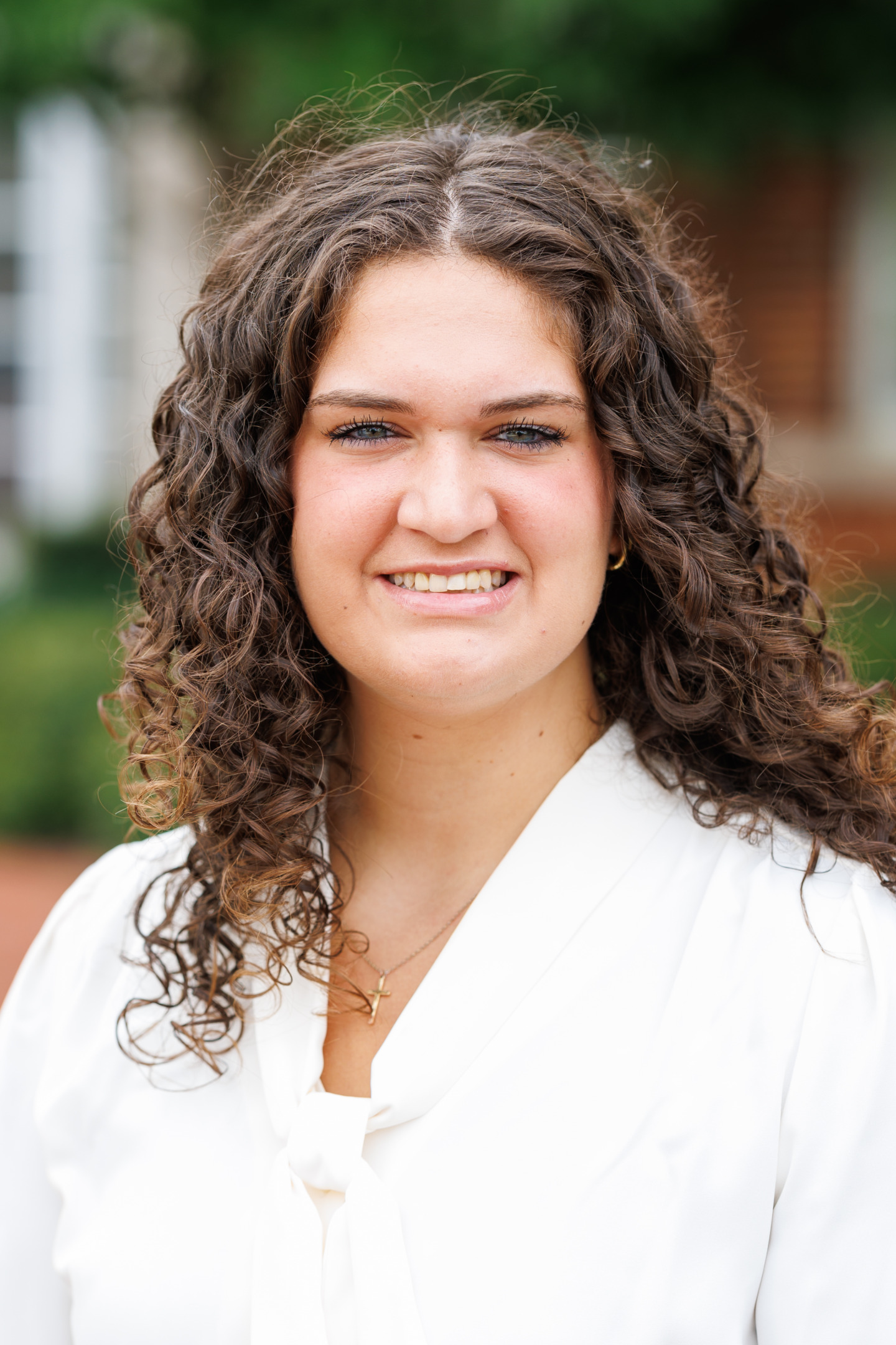 A person with curly brown hair and a white blouse smiles warmly at the camera outdoors.
