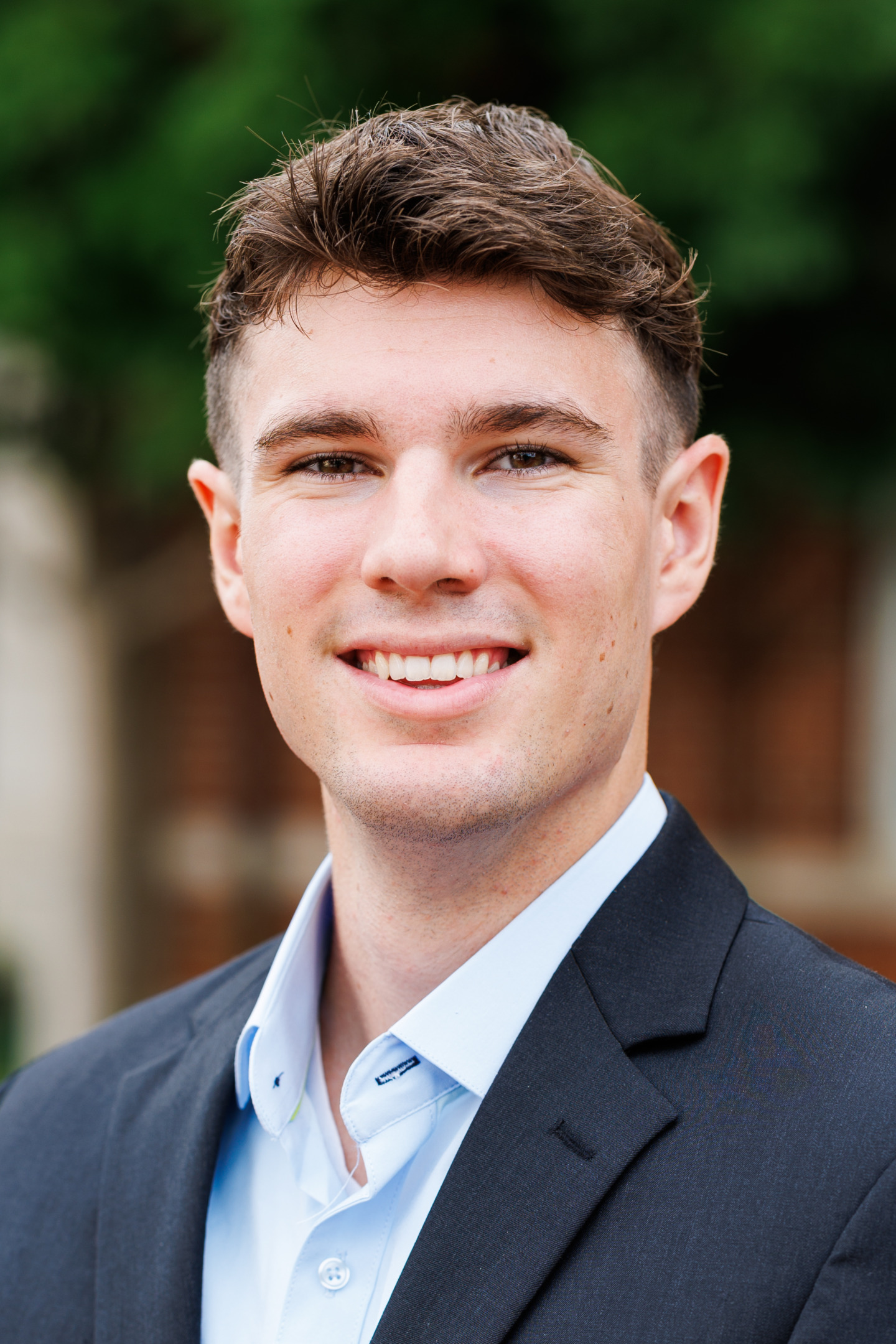 A young person with short brown hair smiles warmly while wearing a dark suit jacket and a light blue collared shirt, set against a softly blurred outdoor background.