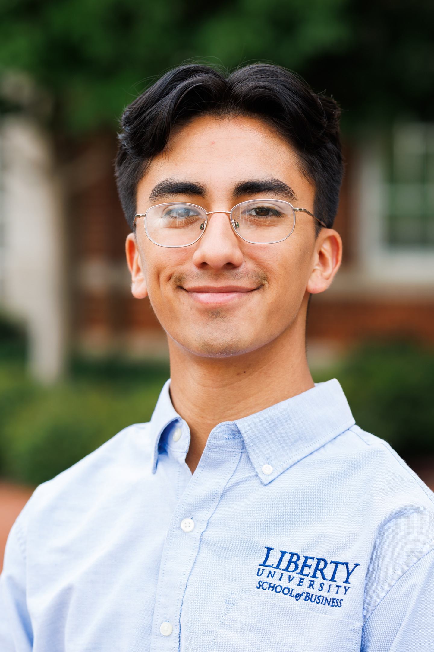 A young person with glasses smiles while wearing a light blue button-down shirt featuring the Liberty University School of Business logo.