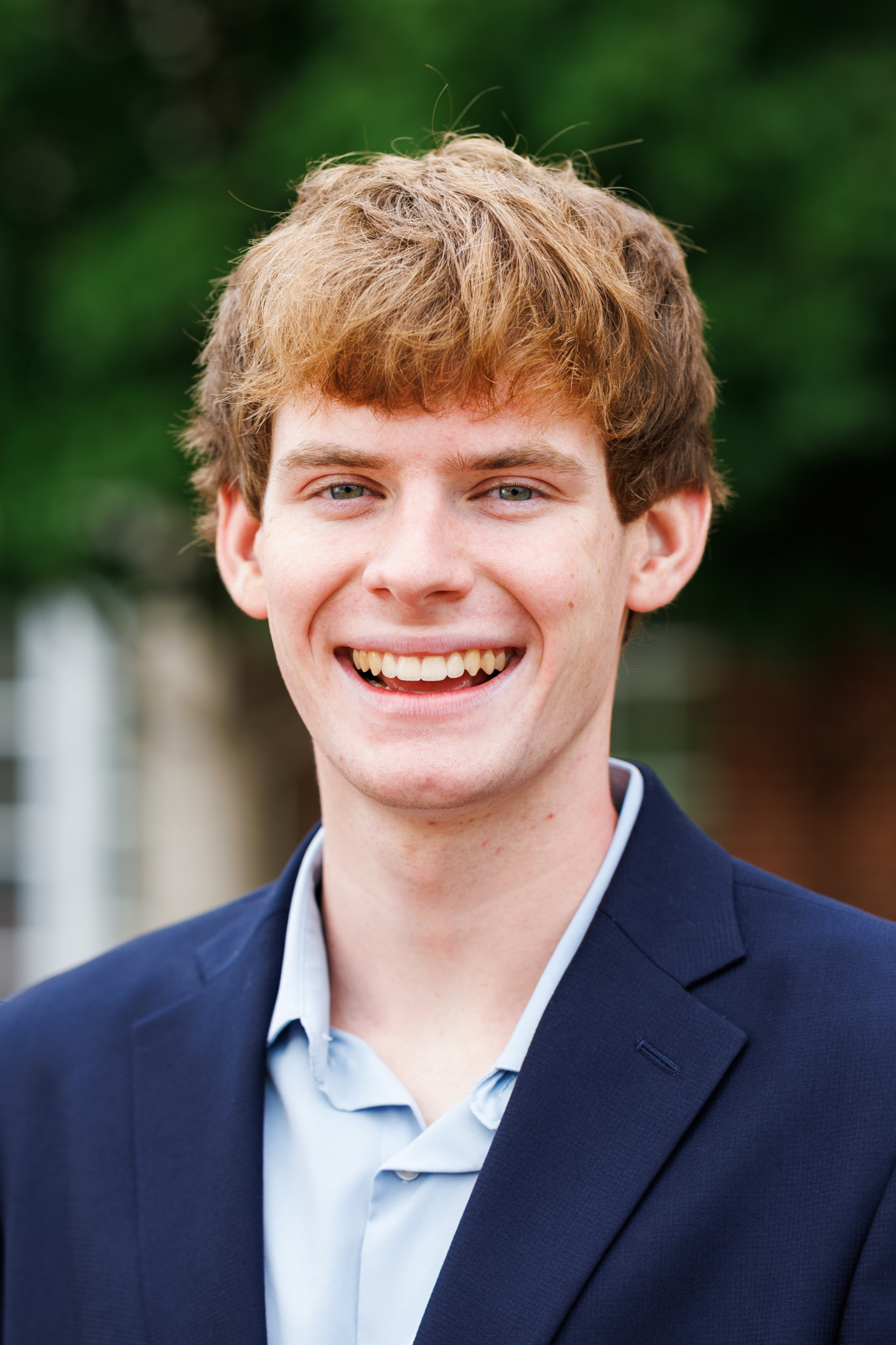 A smiling young person with reddish-brown hair, wearing a navy blazer over a light blue shirt, stands outdoors with a blurred green background.