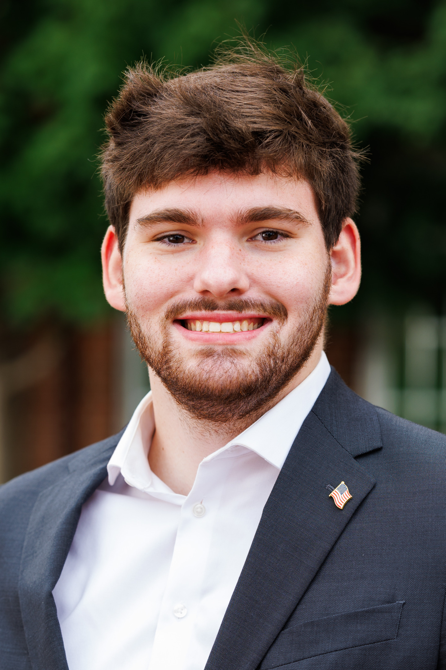 A young person with short brown hair and a beard smiles while wearing a dark suit jacket over a white collared shirt, with a small American flag pin on the lapel.