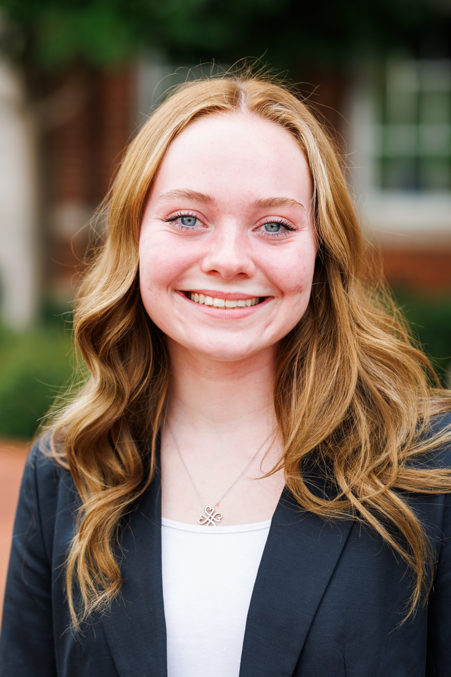 A young person with long, wavy blonde hair smiles warmly while wearing a dark blazer over a white top, standing outdoors with a blurred building background.