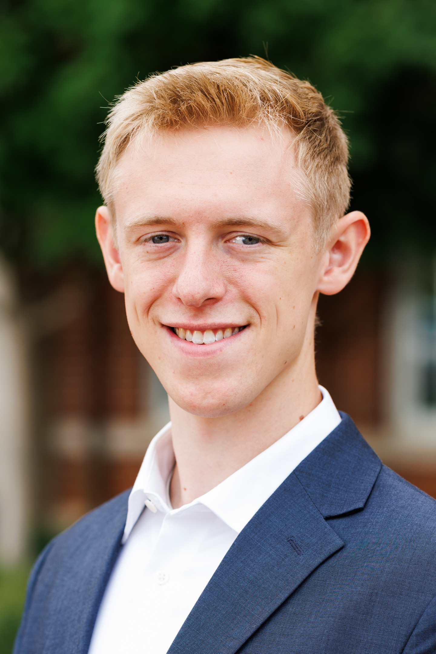 A young person with light hair and a smile, wearing a dark suit jacket and white shirt, stands outdoors with greenery in the background.