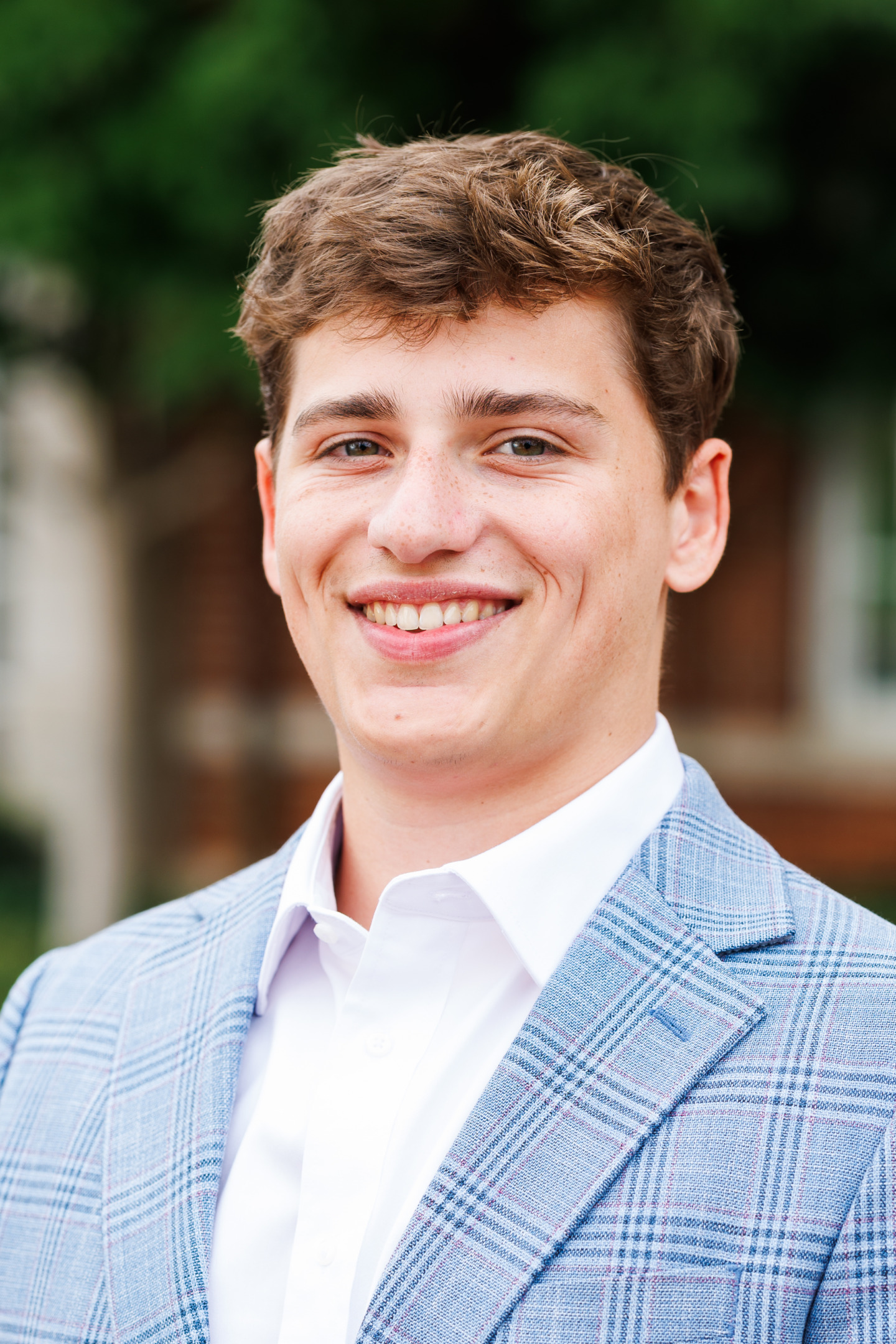A young person with curly brown hair smiles warmly while wearing a light blue plaid blazer over a white collared shirt, set against a blurred outdoor background.