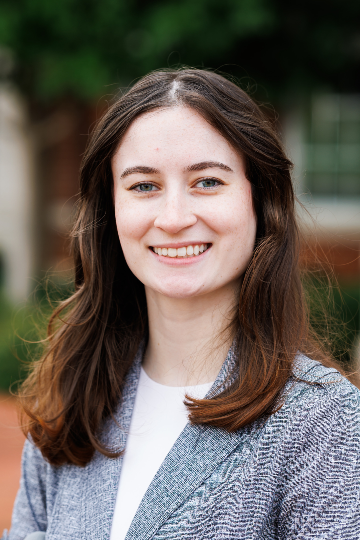 A young person with long brown hair smiles warmly while wearing a gray blazer over a white shirt, set against a softly blurred outdoor background.