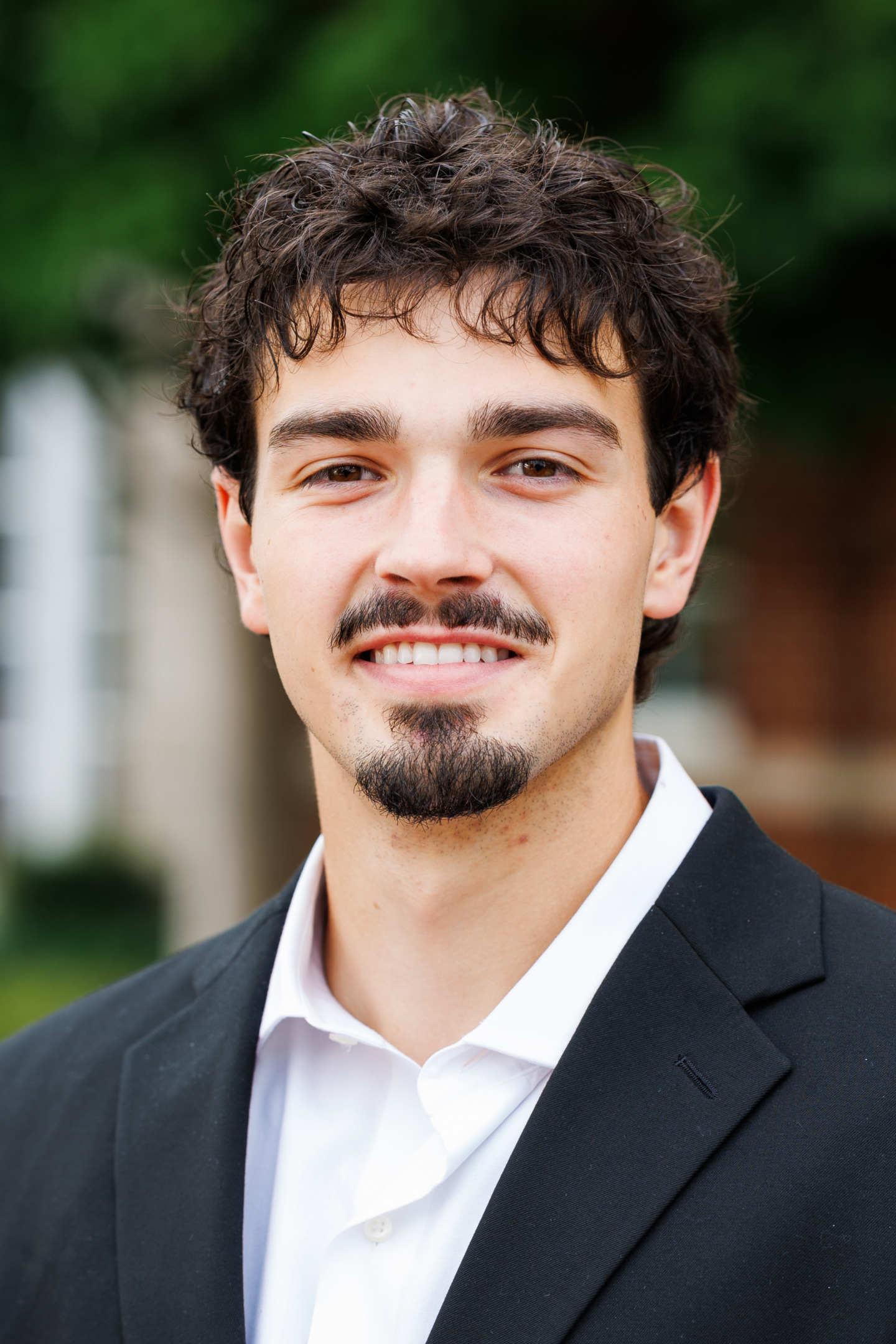 A person with dark curly hair and a goatee smiles while wearing a black suit jacket and white shirt, standing outdoors with greenery in the background.