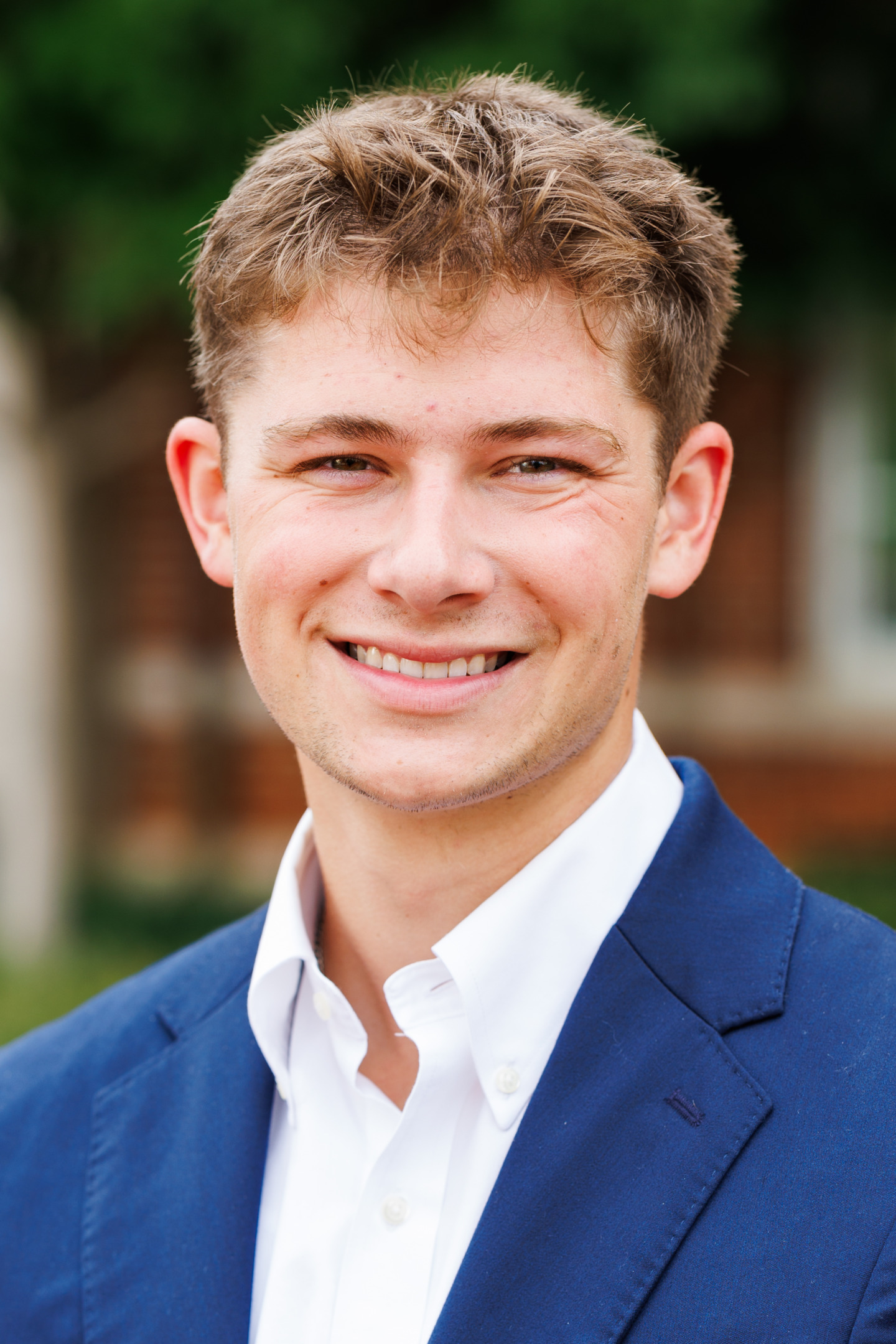A young person with short brown hair smiles warmly while wearing a blue blazer over a white collared shirt, set against a softly blurred outdoor background.