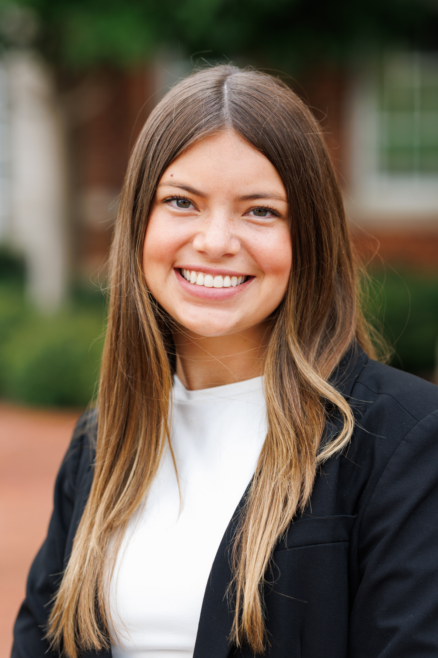 A smiling person with long brown hair, wearing a black blazer over a white top, stands outdoors with a blurred background.