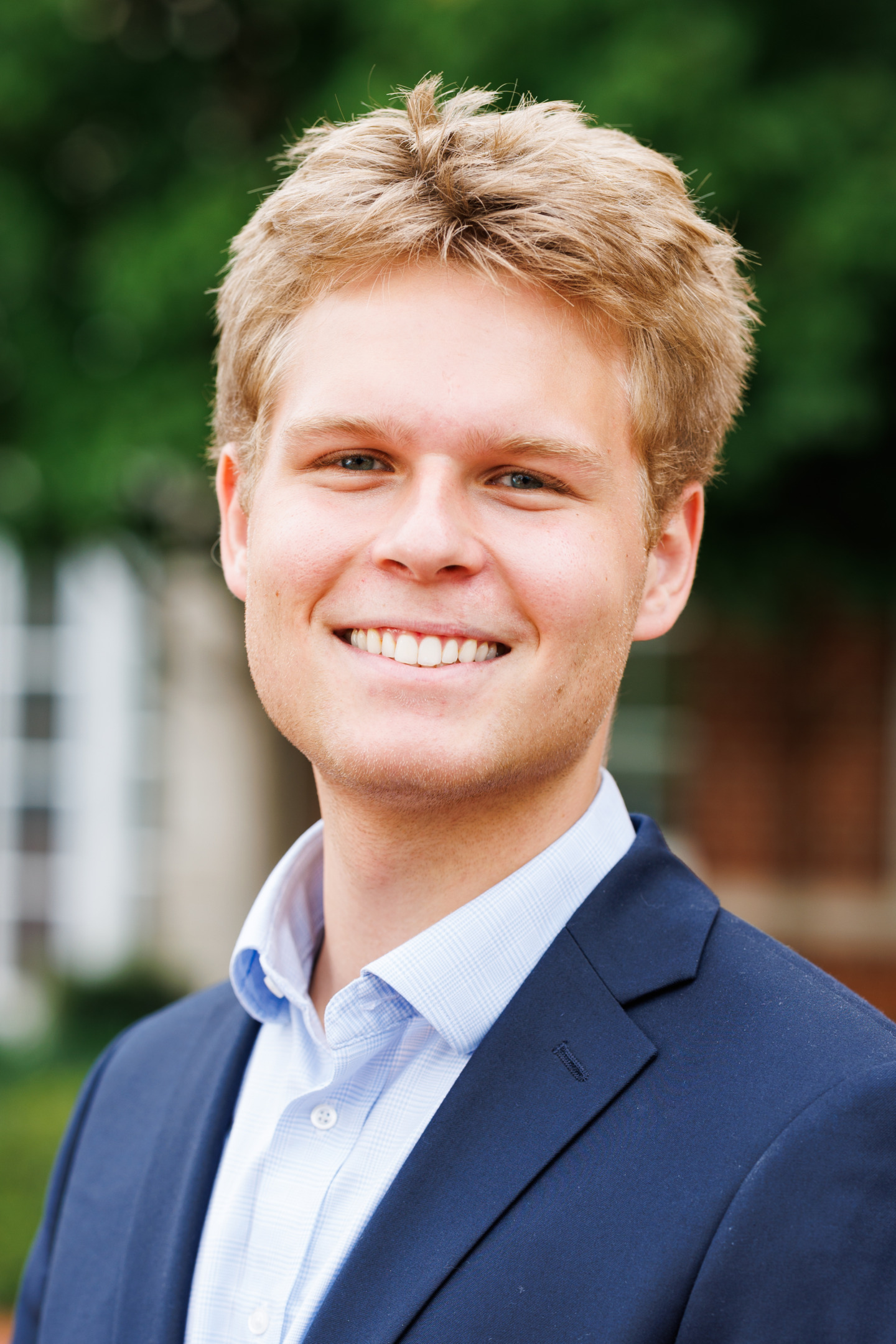 A smiling young person with light-colored hair wears a navy blazer and light blue shirt, standing outdoors with greenery in the background.