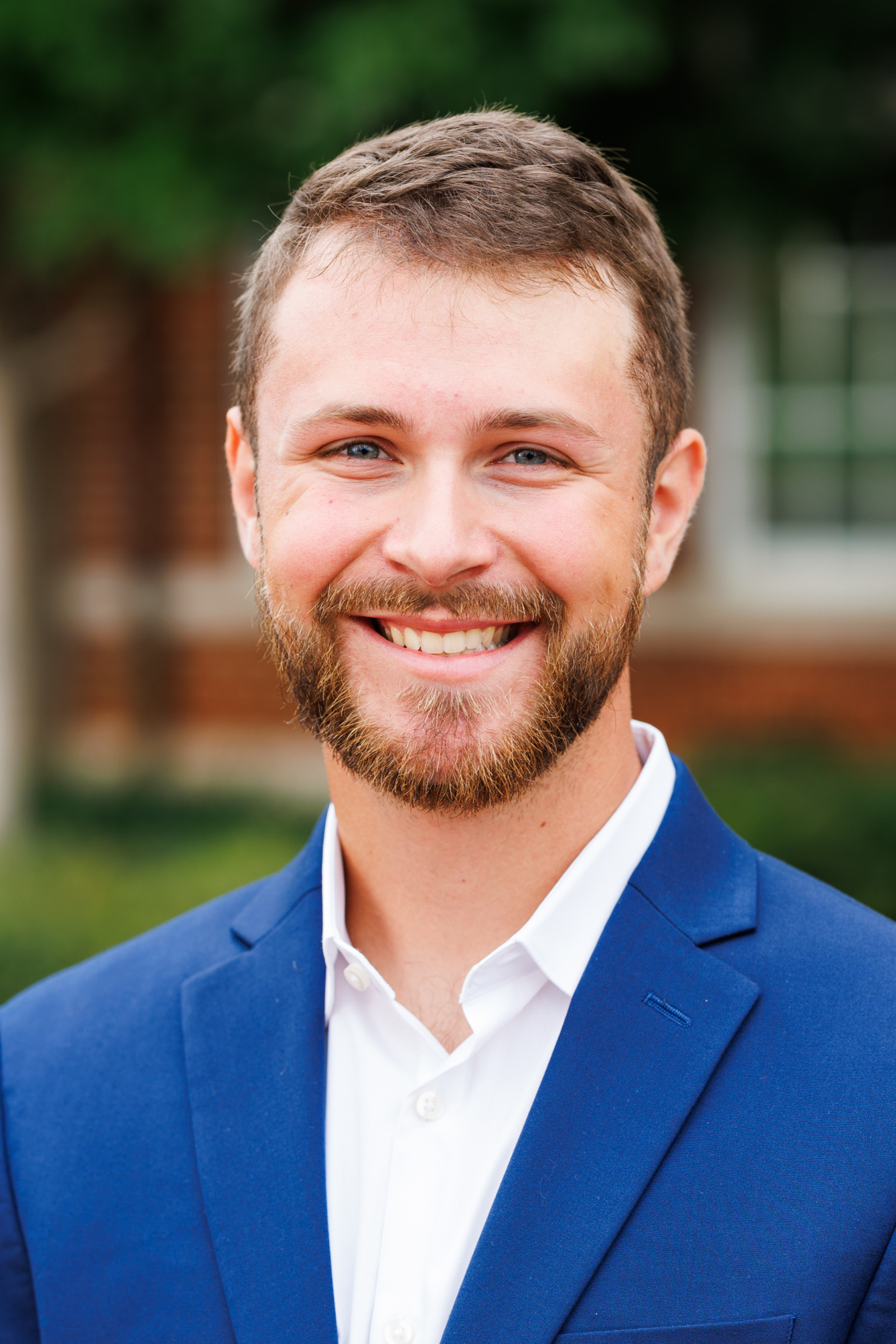 A person with short brown hair and a beard smiles warmly while wearing a blue suit jacket over a white collared shirt, set against a blurred outdoor background.