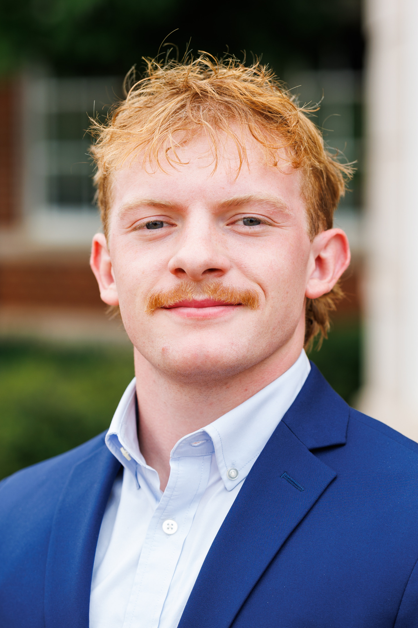 A young person with reddish hair and a mustache smiles while wearing a blue suit jacket over a light blue collared shirt.