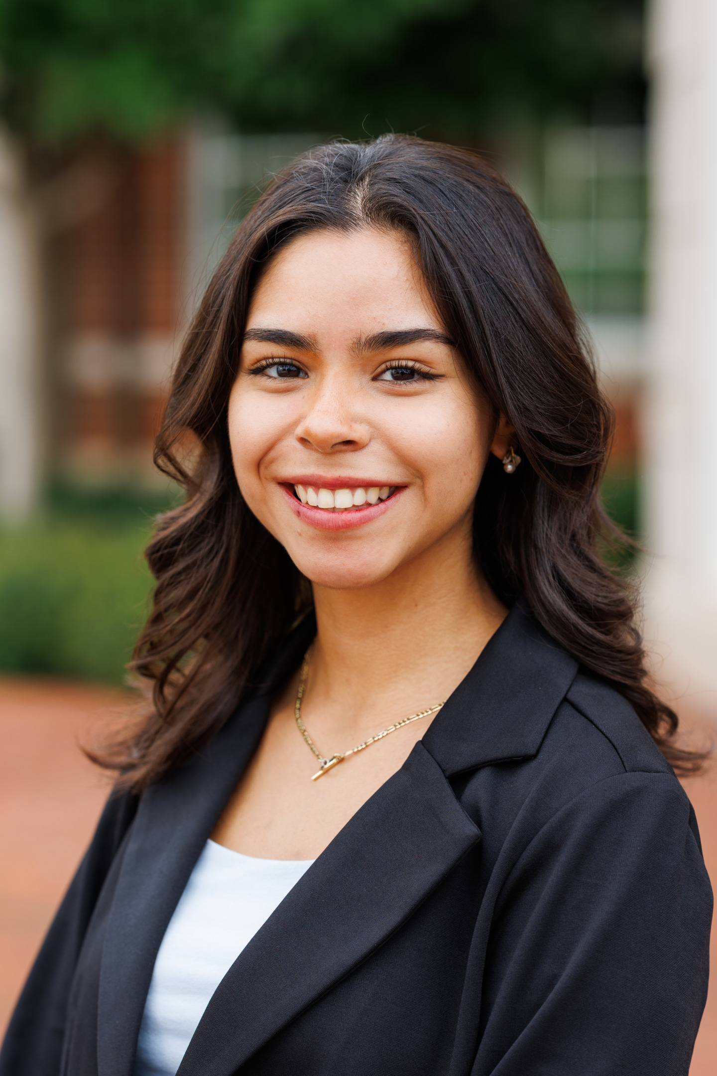 A smiling young person with dark, wavy hair wears a black blazer over a white top, accessorized with a delicate gold necklace and small earrings, standing outdoors with a blurred background.