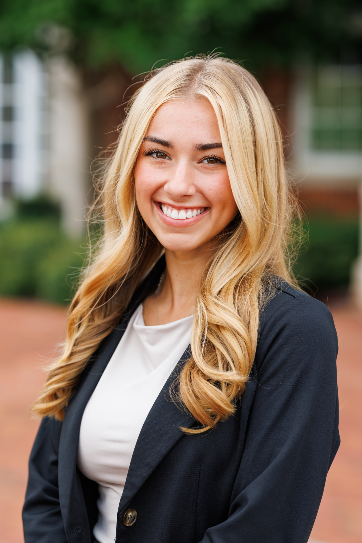 A smiling person with long blonde hair wears a black blazer over a white top, standing outdoors with a blurred background of greenery and buildings.